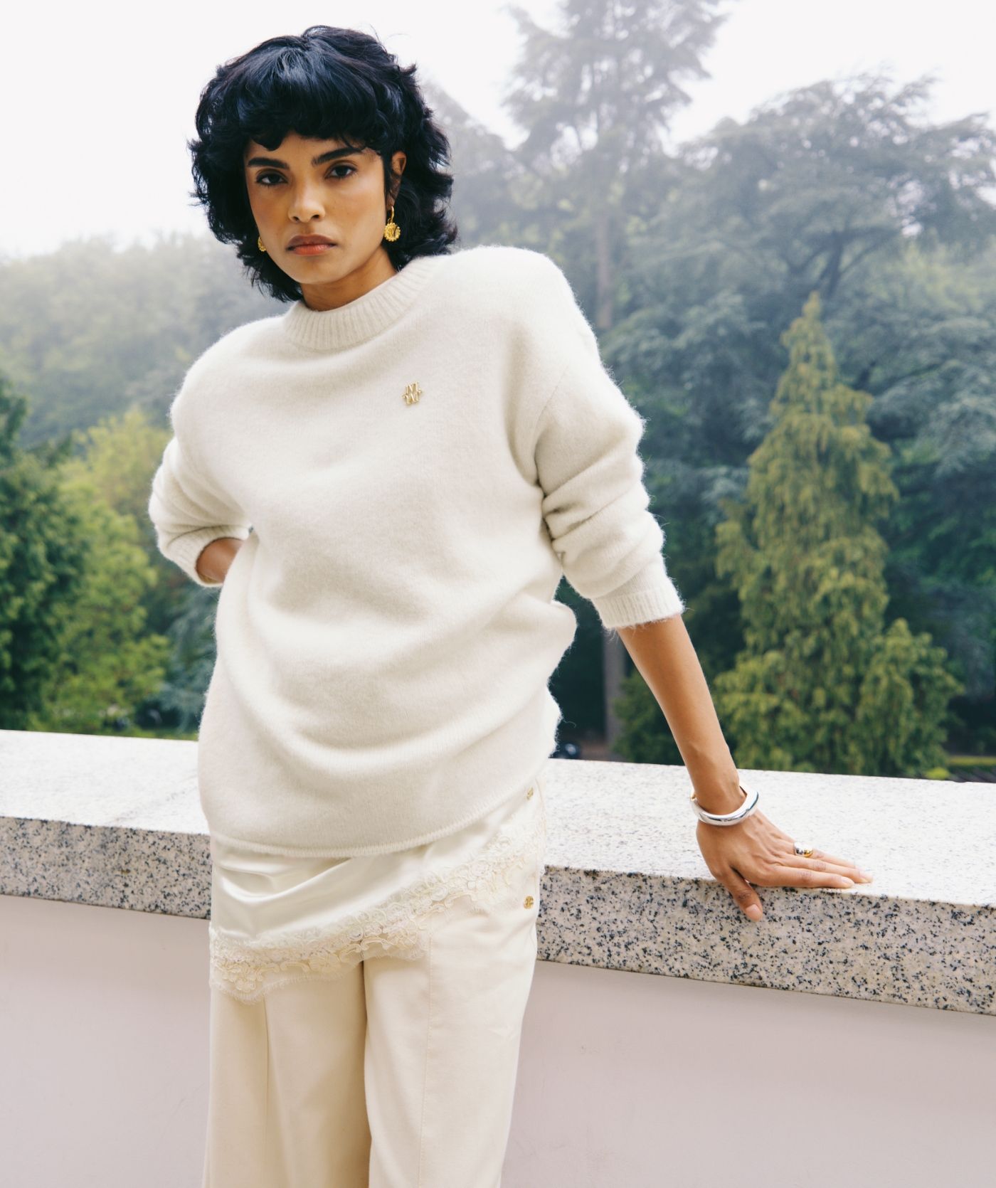 Model draagt oversized witte trui met daaronder een lichtgeel jurken met kanten details aan de boven en onderkant, gecombineerd met beige broek.