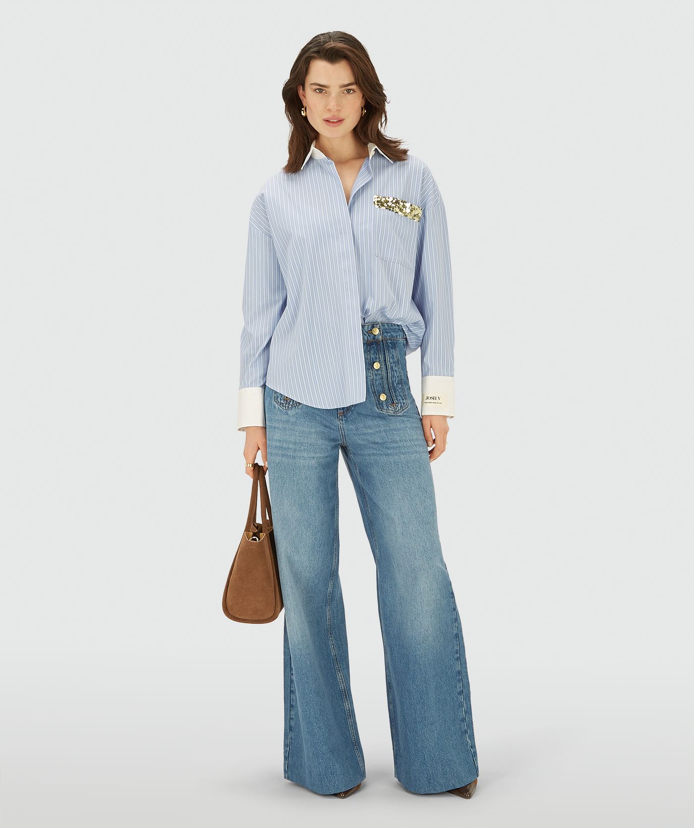 Model draagt een longsleeve oversized  blouse in de kleur Powder Blue Stripe. Ook heeft de blouse een merklabel op de linkermouw en een borstzak afgewerkt met groen-gouden pailletten