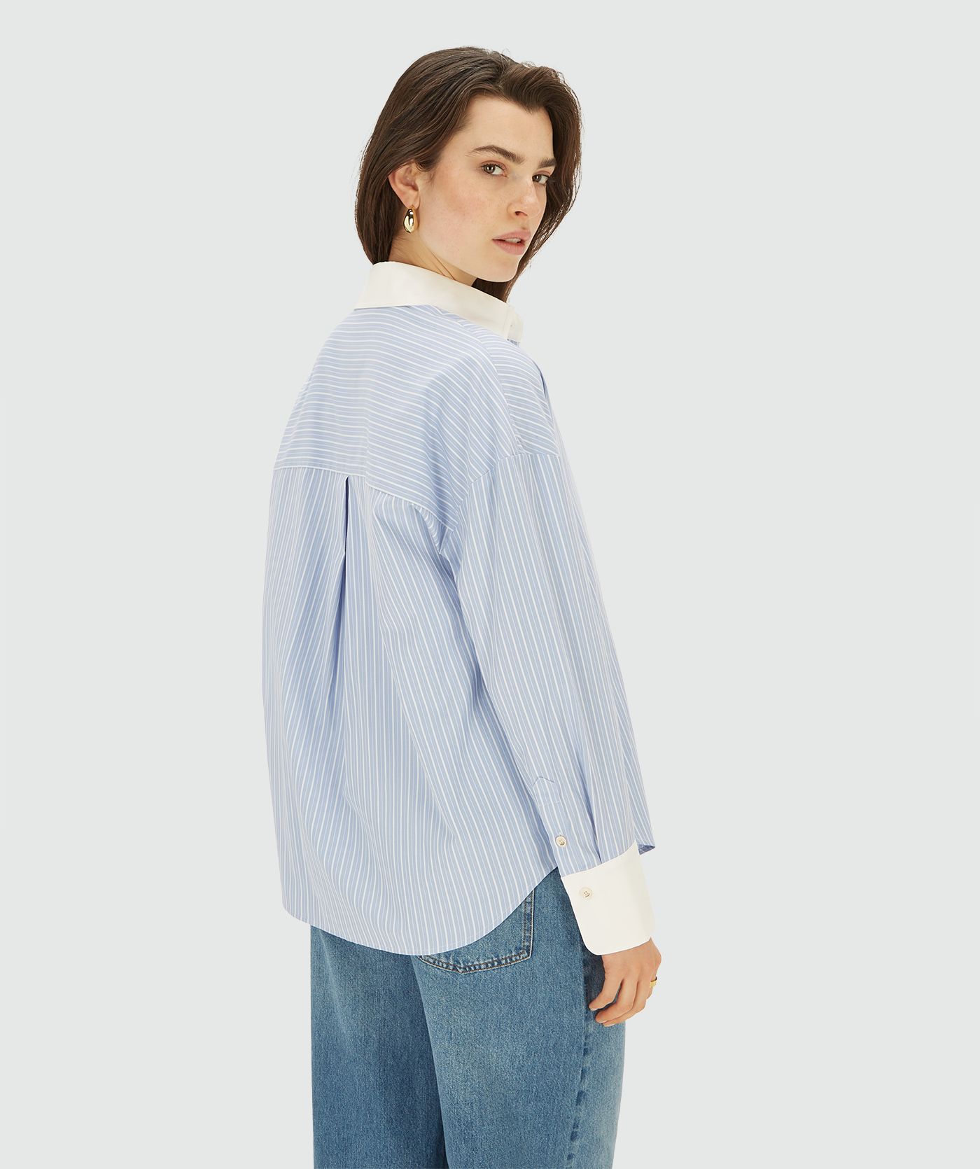 Achteraanzicht van model in een longsleeve oversized  blouse in de kleur Powder Blue Stripe. Ook heeft de blouse een merklabel op de linkermouw en een borstzak afgewerkt met groen-gouden pailletten