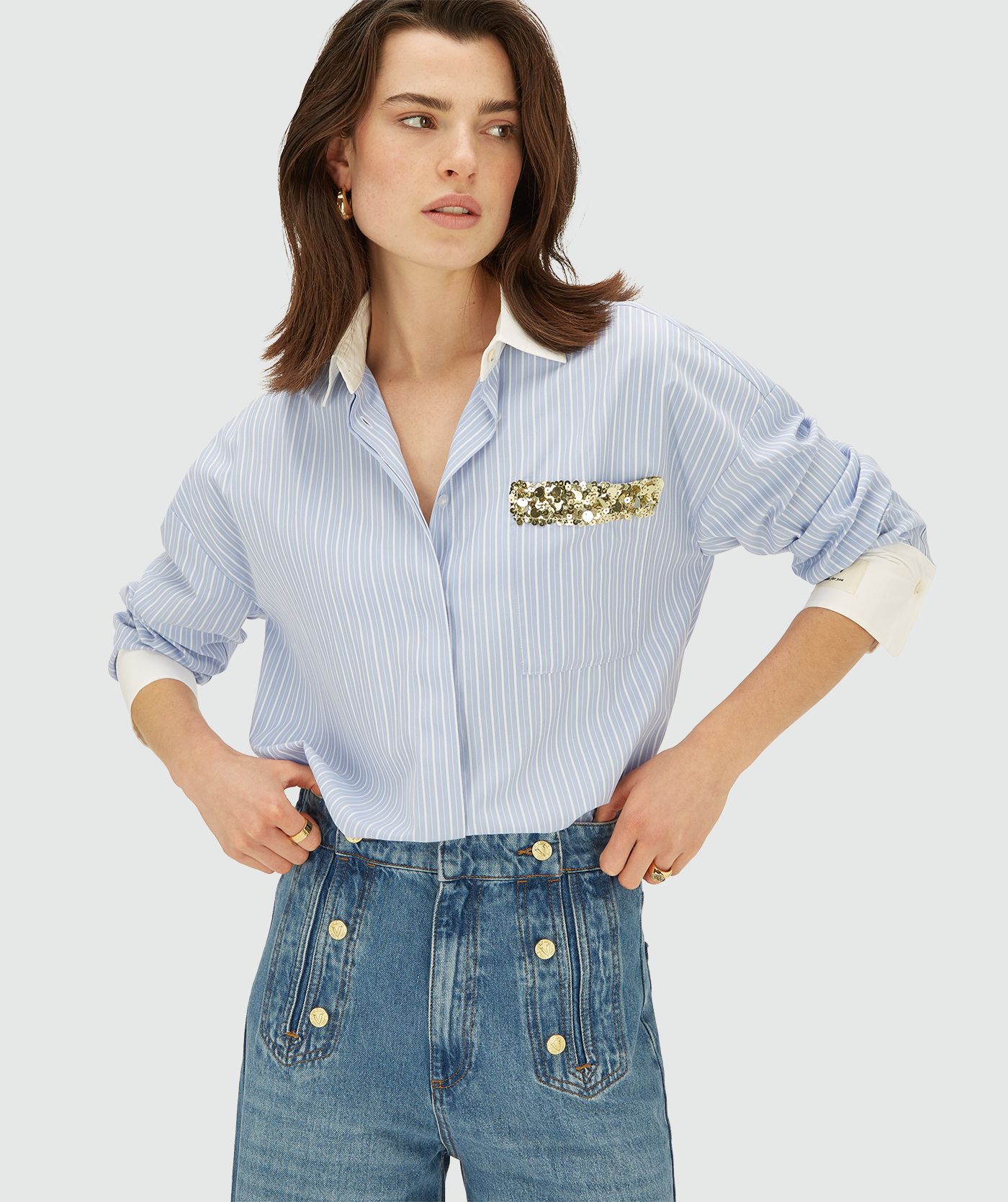 Model draagt een longsleeve oversized  blouse in de kleur Powder Blue Stripe. Ook heeft de blouse een merklabel op de linkermouw en een borstzak afgewerkt met groen-gouden pailletten