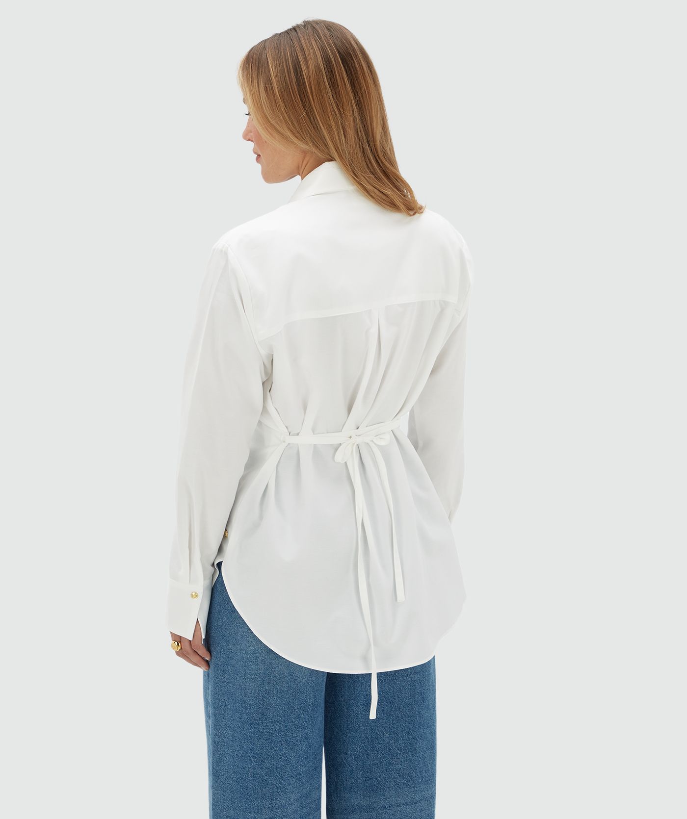 Model draagt een witte blouse aan de bovenkant vier gouden knopen. De blouse is gecombineerd op een jeans.