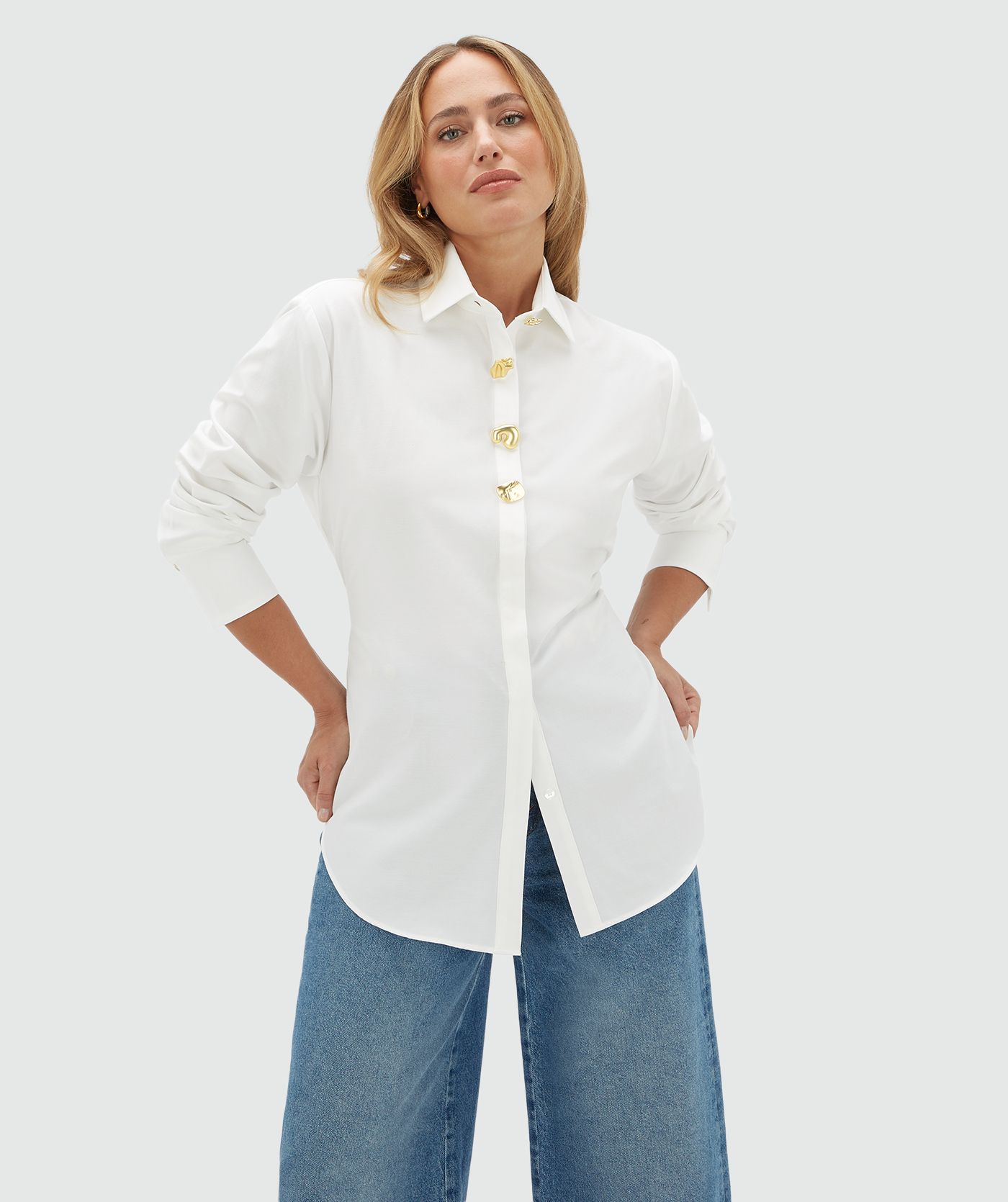 Model draagt een witte blouse aan de bovenkant vier gouden knopen. De blouse is gecombineerd op een jeans.