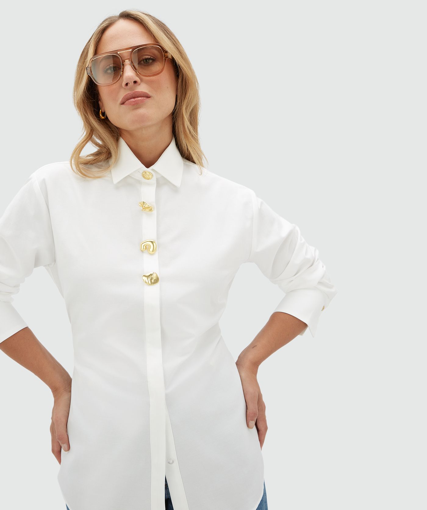 Model draagt een witte blouse aan de bovenkant vier gouden knopen. De blouse is gecombineerd op een jeans.