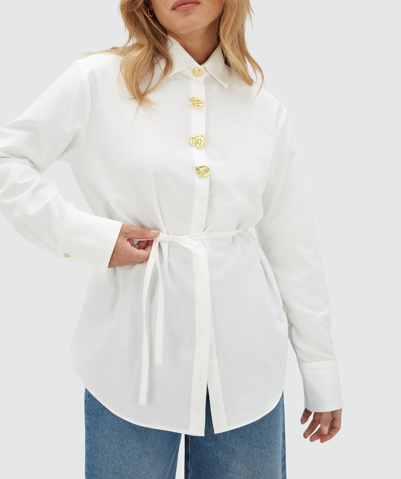 Model draagt een witte blouse aan de bovenkant vier gouden knopen. De blouse is gecombineerd op een jeans.