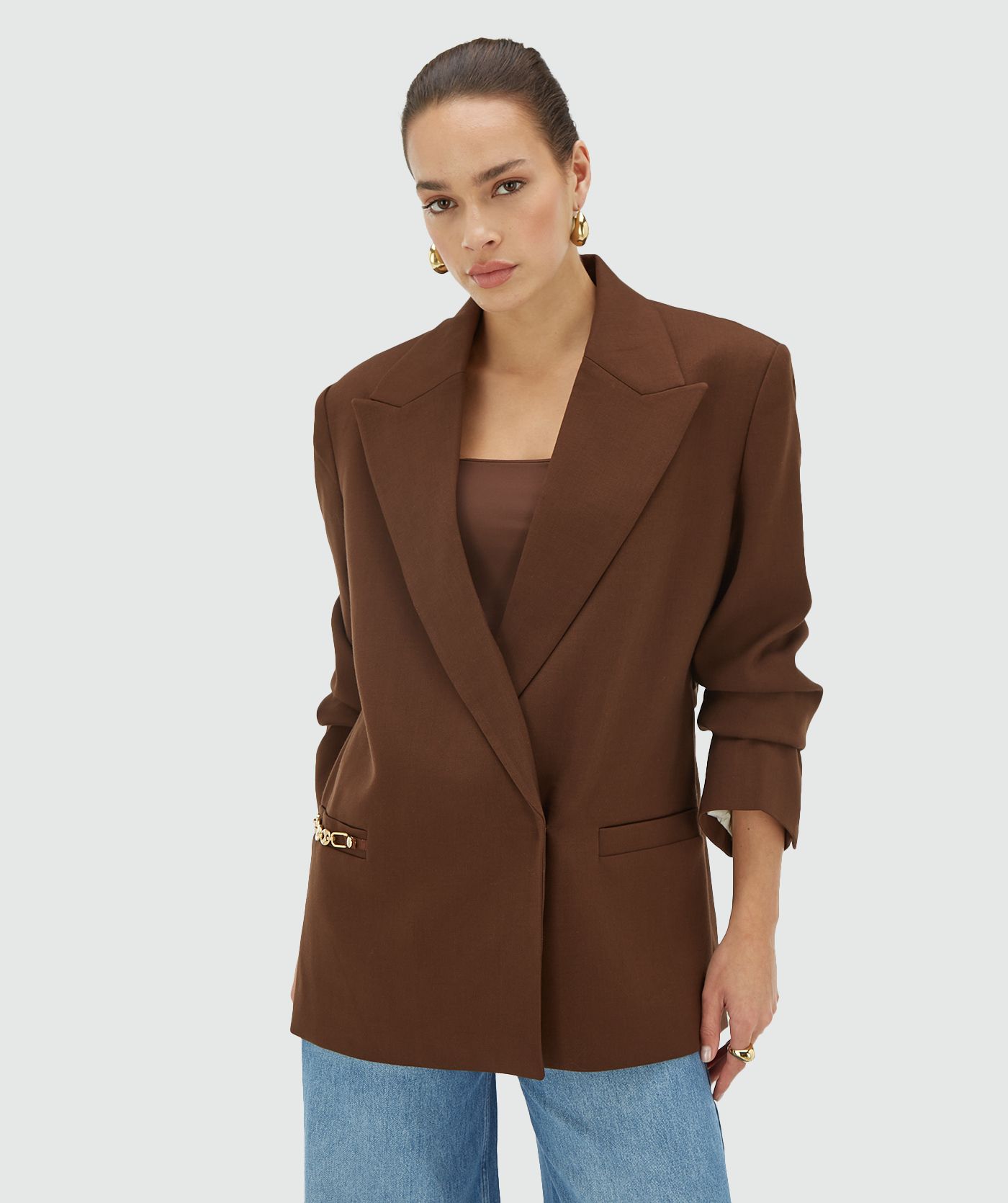 Model draagt een oversized Dark Brown blazer met schoudervullingen en is voorzien van een JOSH V branded chain op de steekzak en een riem met JOSH V branded haakjes - Josh V