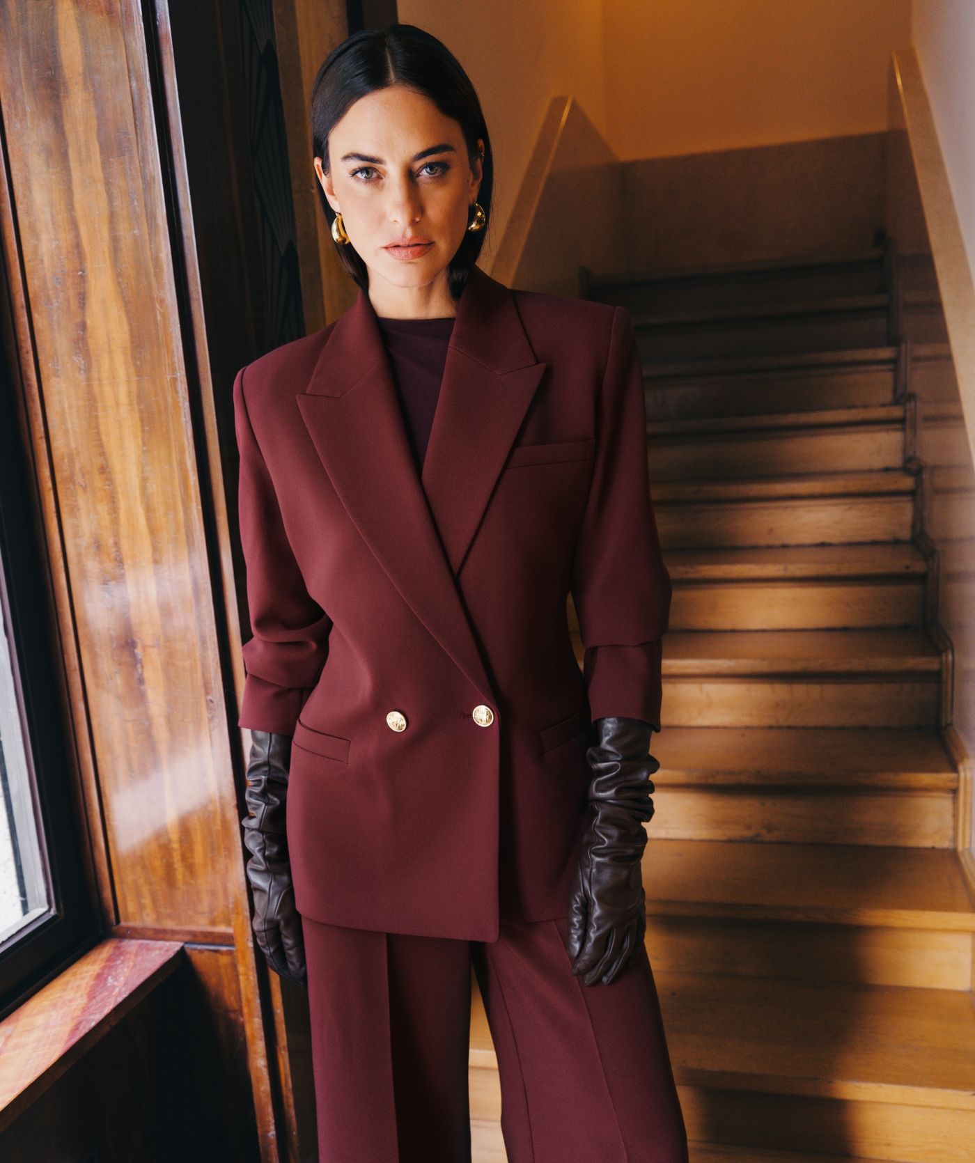 Model staat bij een trap en draagt de donkerrode blazer gecombineerd met de bijpassende pantalon en lange handschoenen. 
