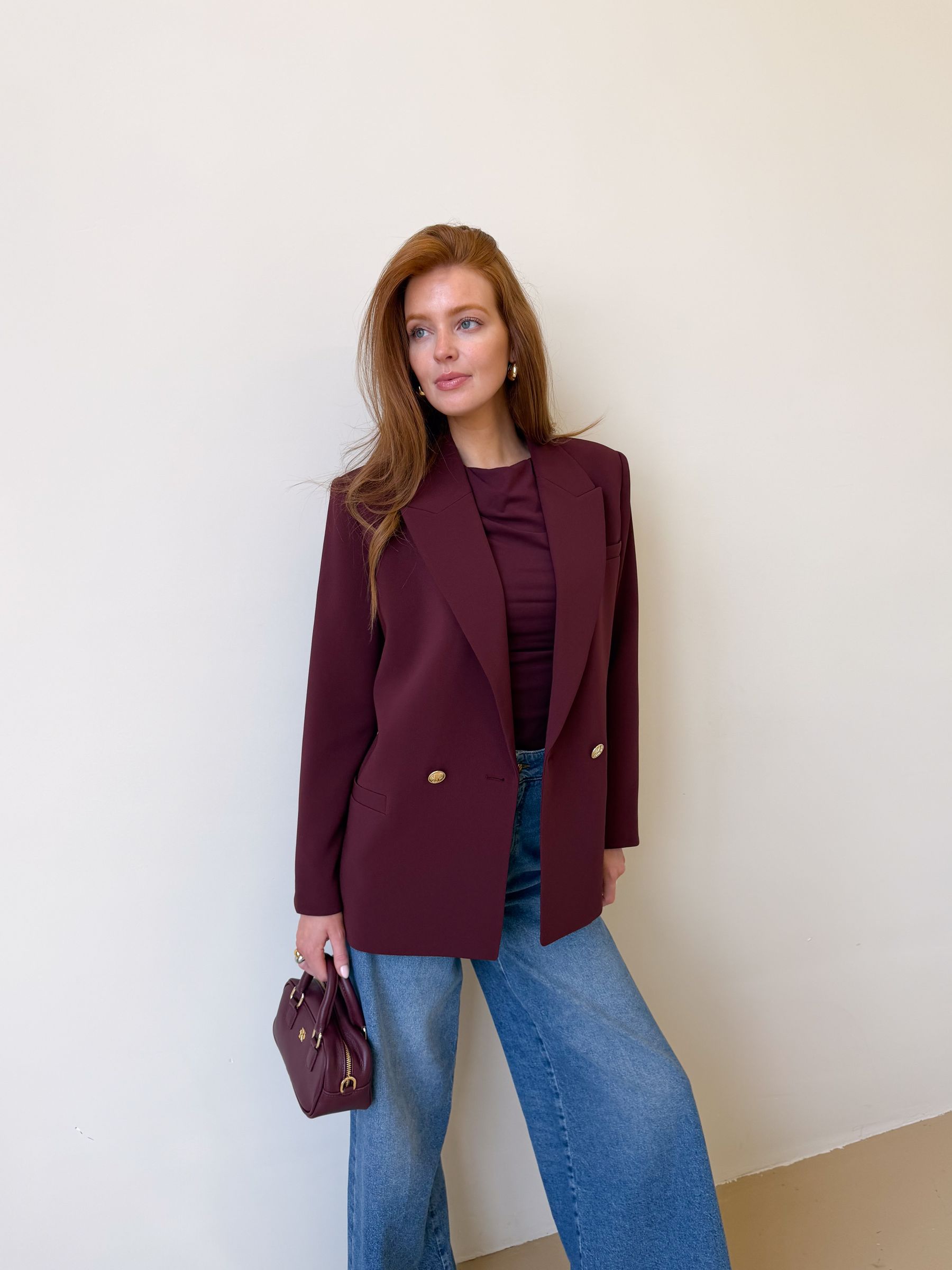 Model draagt een donkerrode blazer met een riempje achter zodat er meer taille gecreëerd kan worden. De blazer is gecombineerd met een jeans.