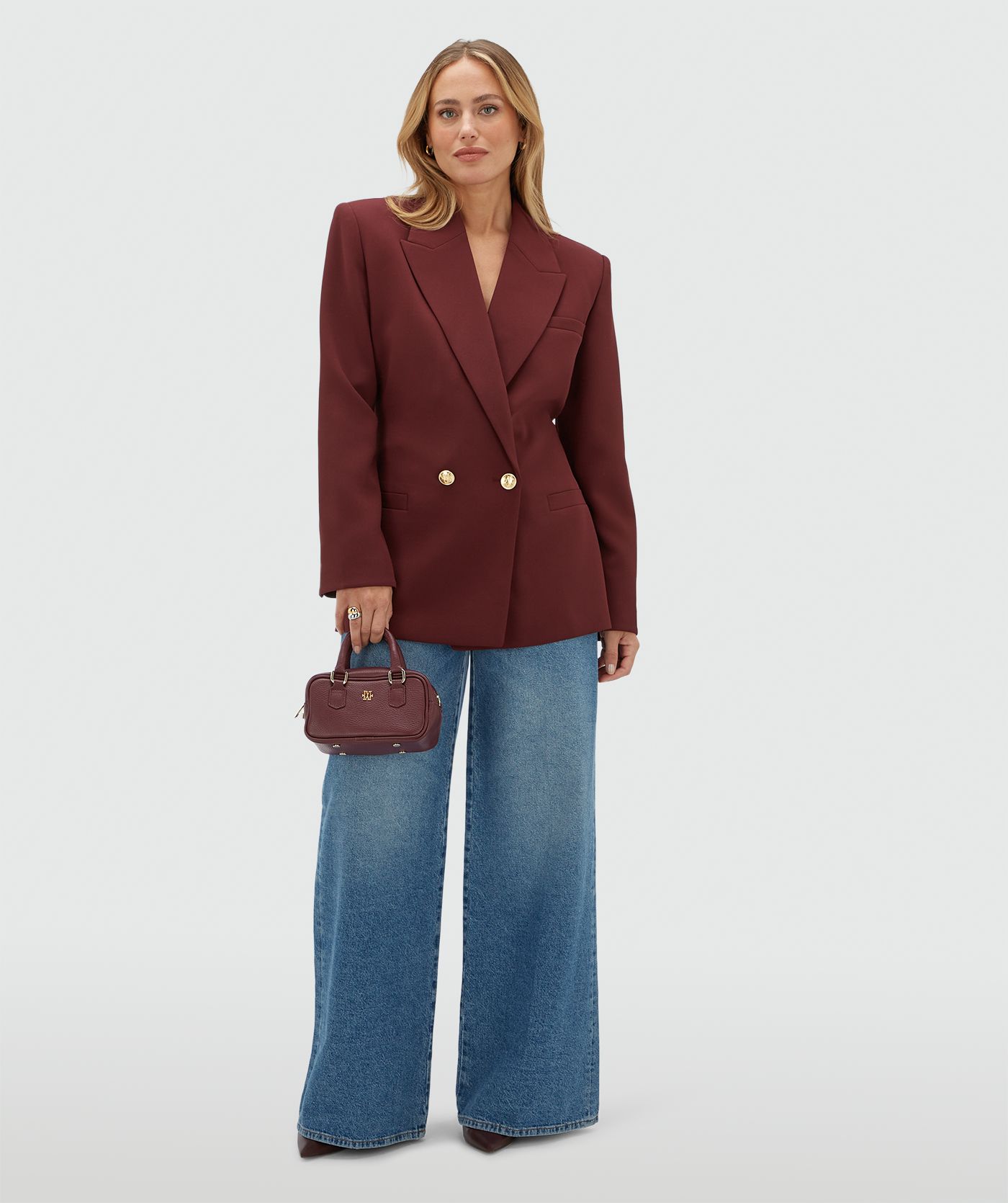 Model draagt een donkerrode blazer met een riempje achter zodat er meer taille gecreëerd kan worden en heeft gouden knopen. De blazer is gecombineerd met een jeans.