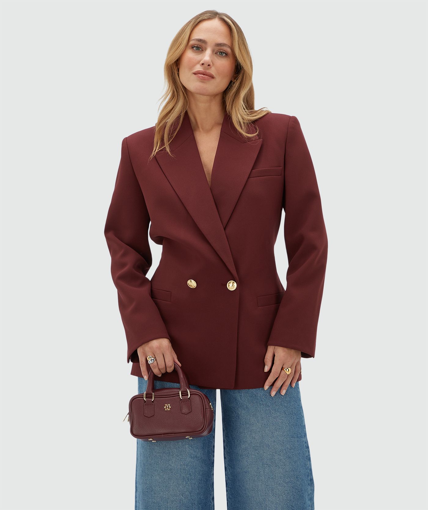 Model draagt een donkerrode blazer met een riempje achter zodat er meer taille gecreëerd kan worden en heeft gouden knopen. De blazer is gecombineerd met een jeans.