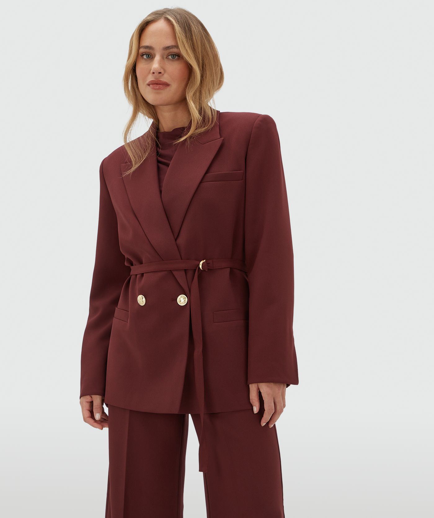 Model draagt een donkerrode blazer met een riempje. De blazer is gecombineerd met de bijpassende pantalon.