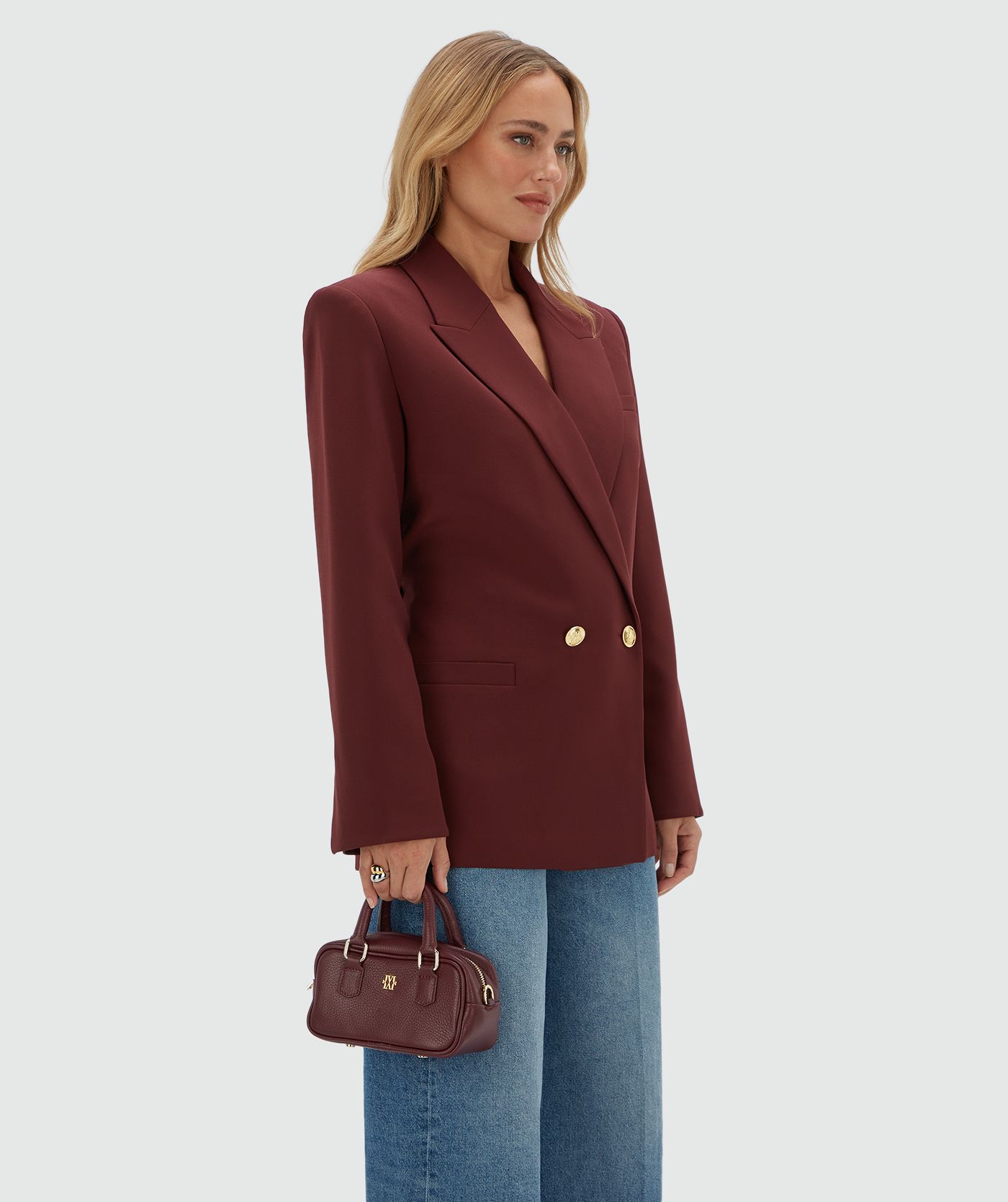Model draagt een donkerrode blazer met een riempje achter zodat er meer taille gecreëerd kan worden en heeft gouden knopen. De blazer is gecombineerd met een jeans.