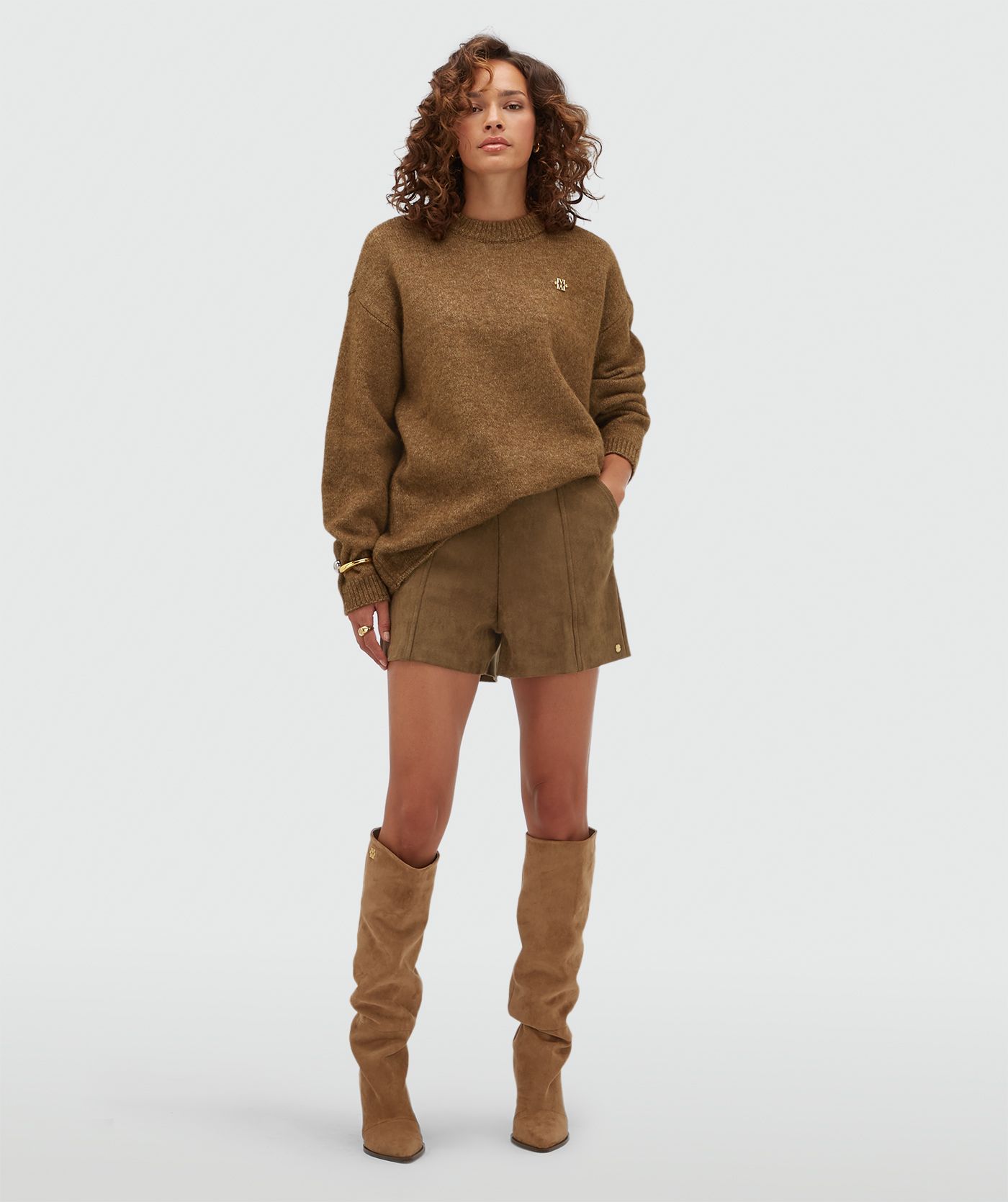 Model draagt een donkerbruine oversized trui gecombineerd met een kort suede broekje.