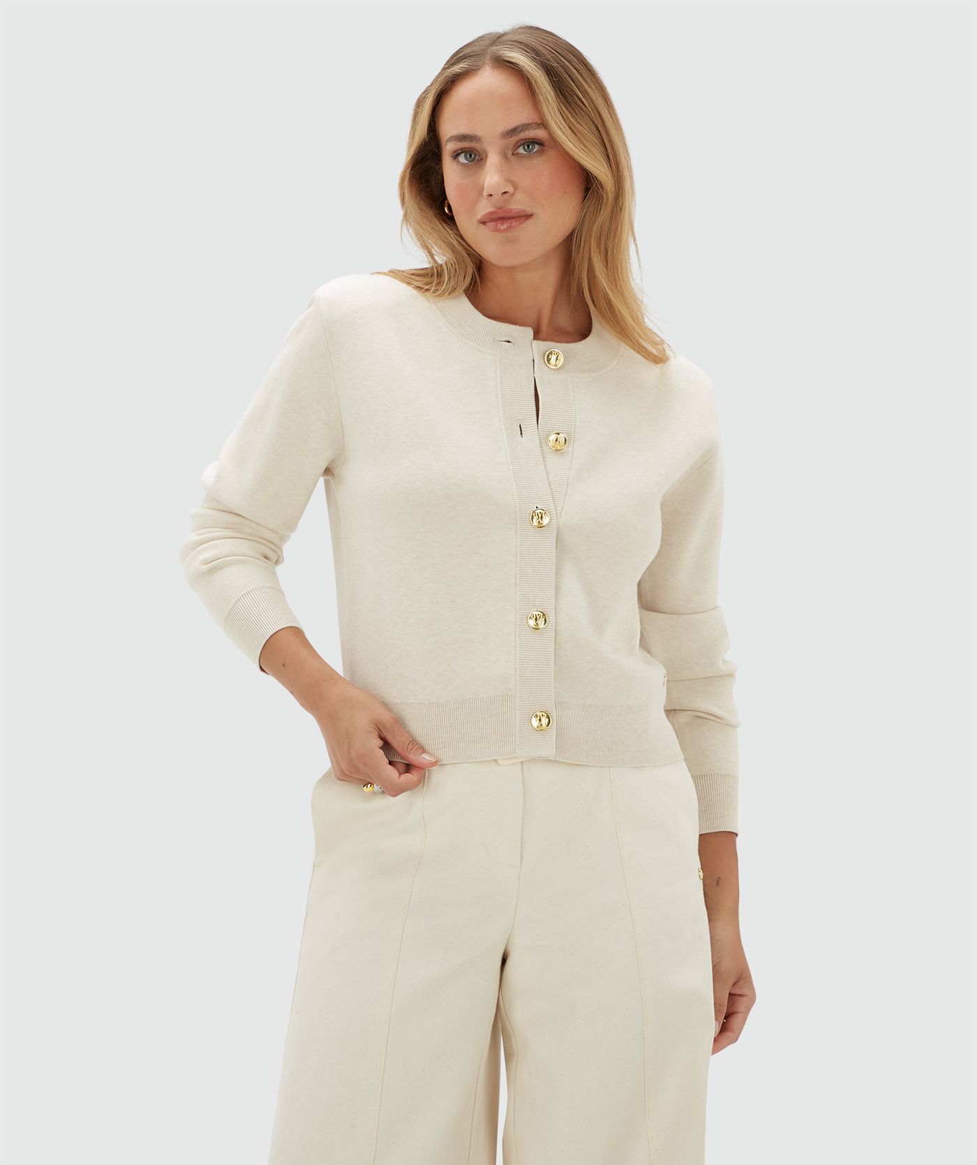 Model draagt een gebroken wit vest met gouden knopen en schoudervullingen op een beige broek.