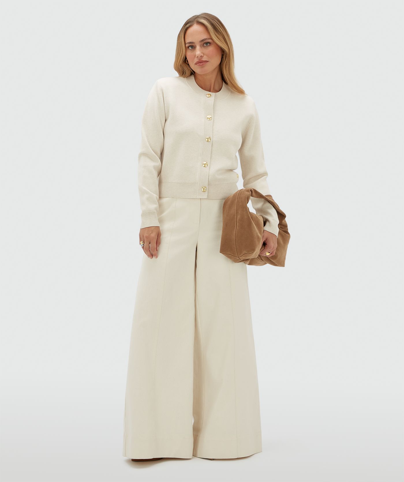 Model draagt een gebroken wit vest met gouden knopen en schoudervullingen op een beige broek.