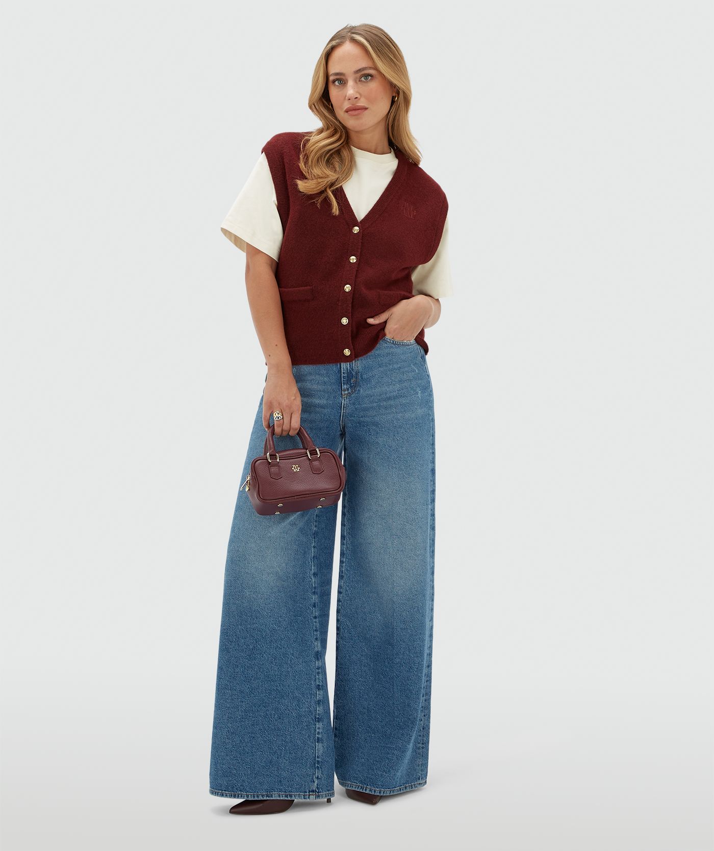 Model draagt een donkerrood gilet met gouden knoopjes en een JOSH V logo op de borst. Het gilet is gecombineerd met een wit T-shirt en een jeans.