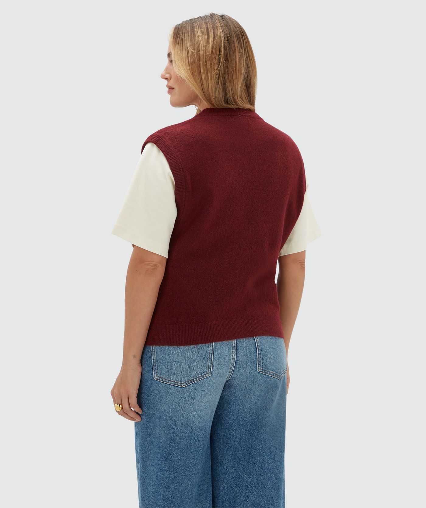 Achteraanzicht van een model in een donkerrood gilet gecombineerd met een wit T-shirt en een jeans.