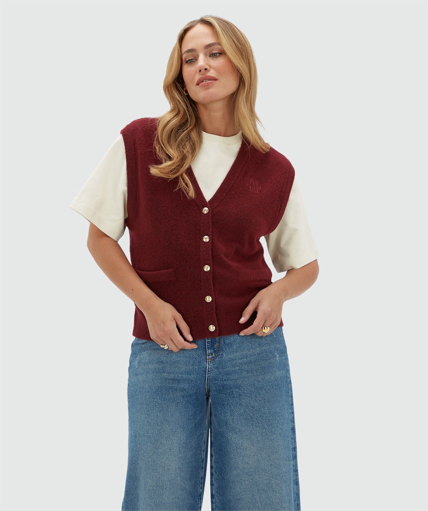 Model draagt een donkerrood gilet met gouden knoopjes en een JOSH V logo op de borst. Het gilet is gecombineerd met een wit T-shirt en een jeans.