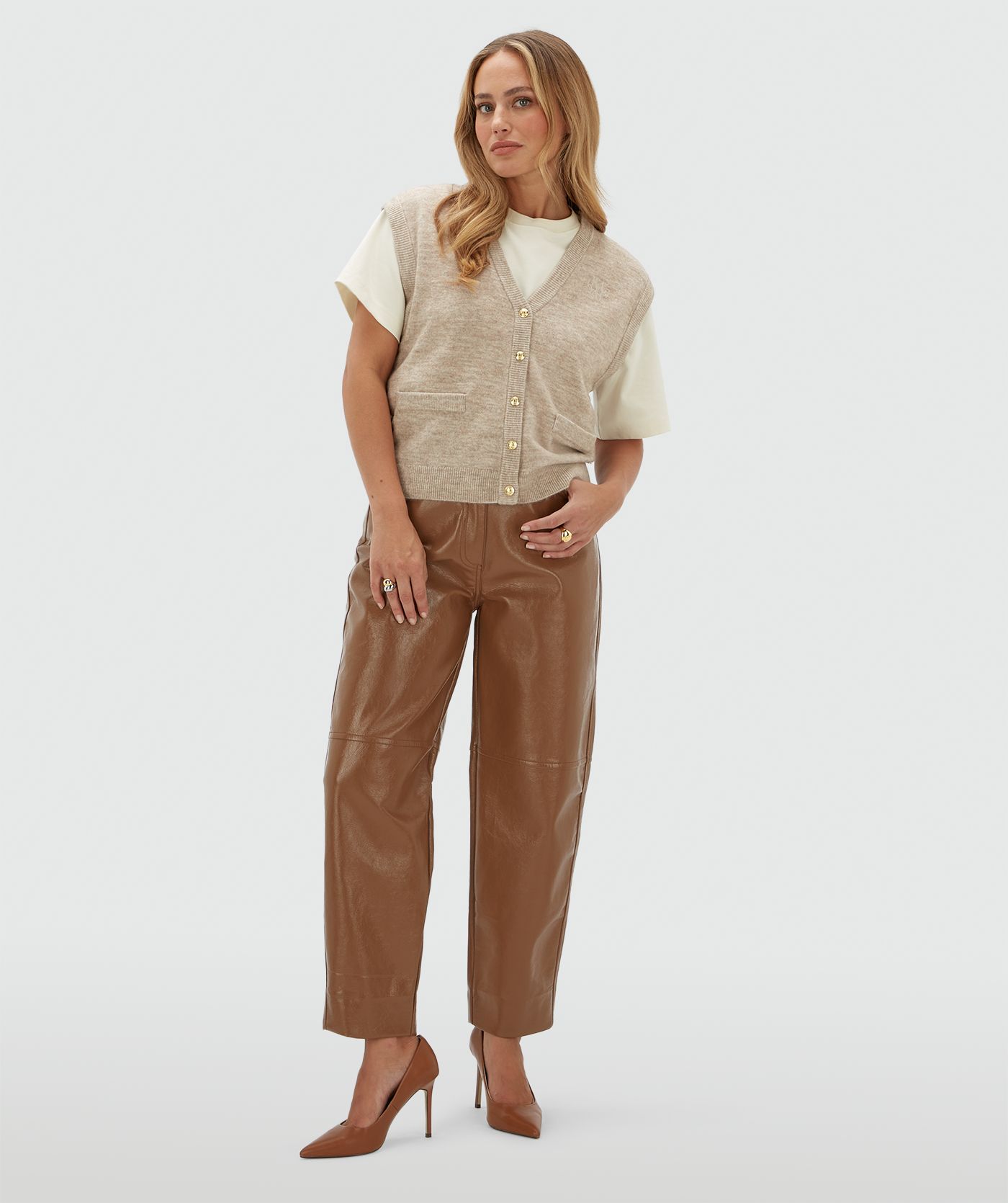 Model draagt een beige gilet met gouden knoopjes en een JOSH V logo op de borst. Het gilet is gecombineerd met een wit T-shirt en een bruine lak look broek.