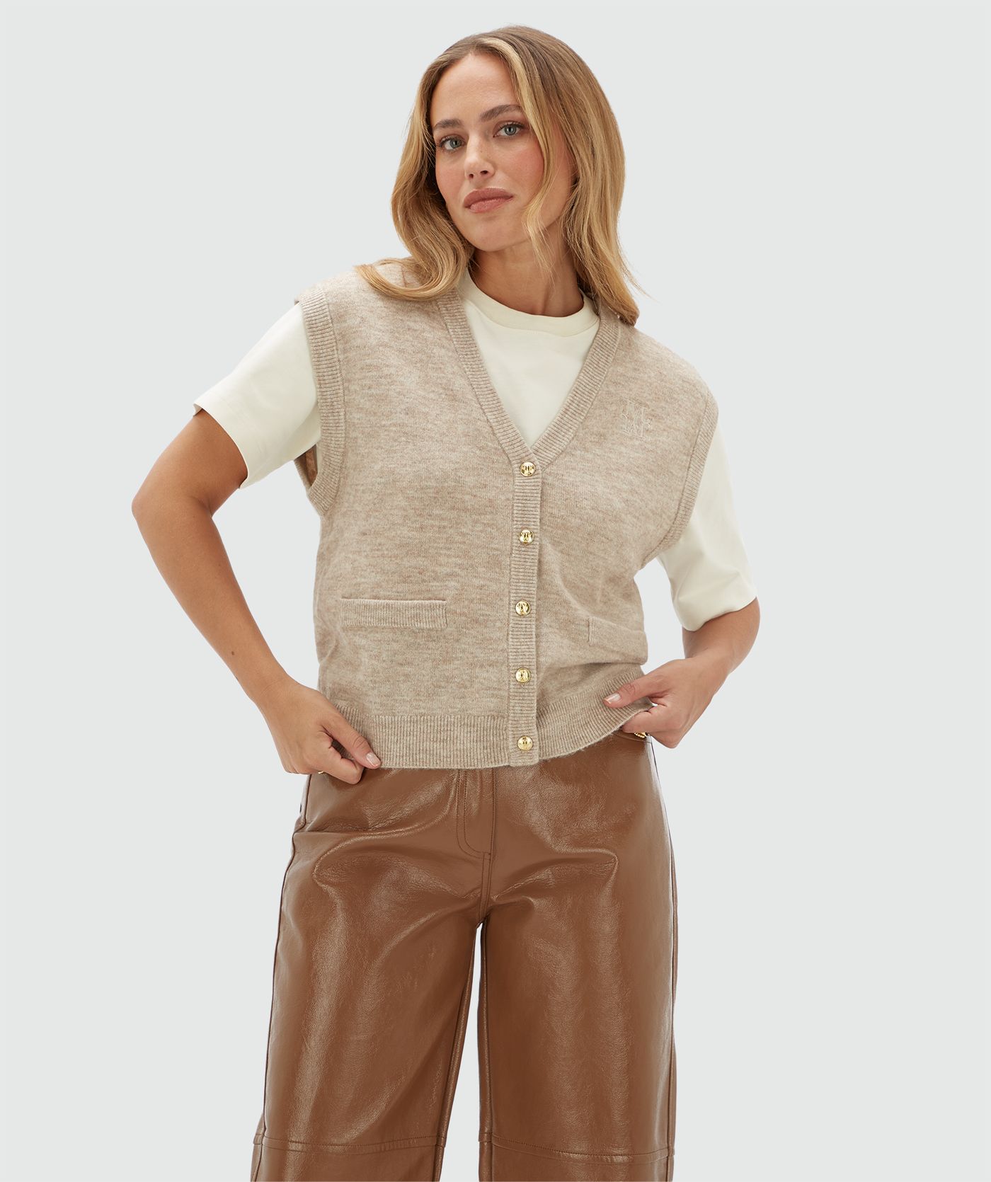 Model draagt een beige gilet met gouden knoopjes en een JOSH V logo op de borst. Het gilet is gecombineerd met een wit T-shirt en een bruine lak look broek.