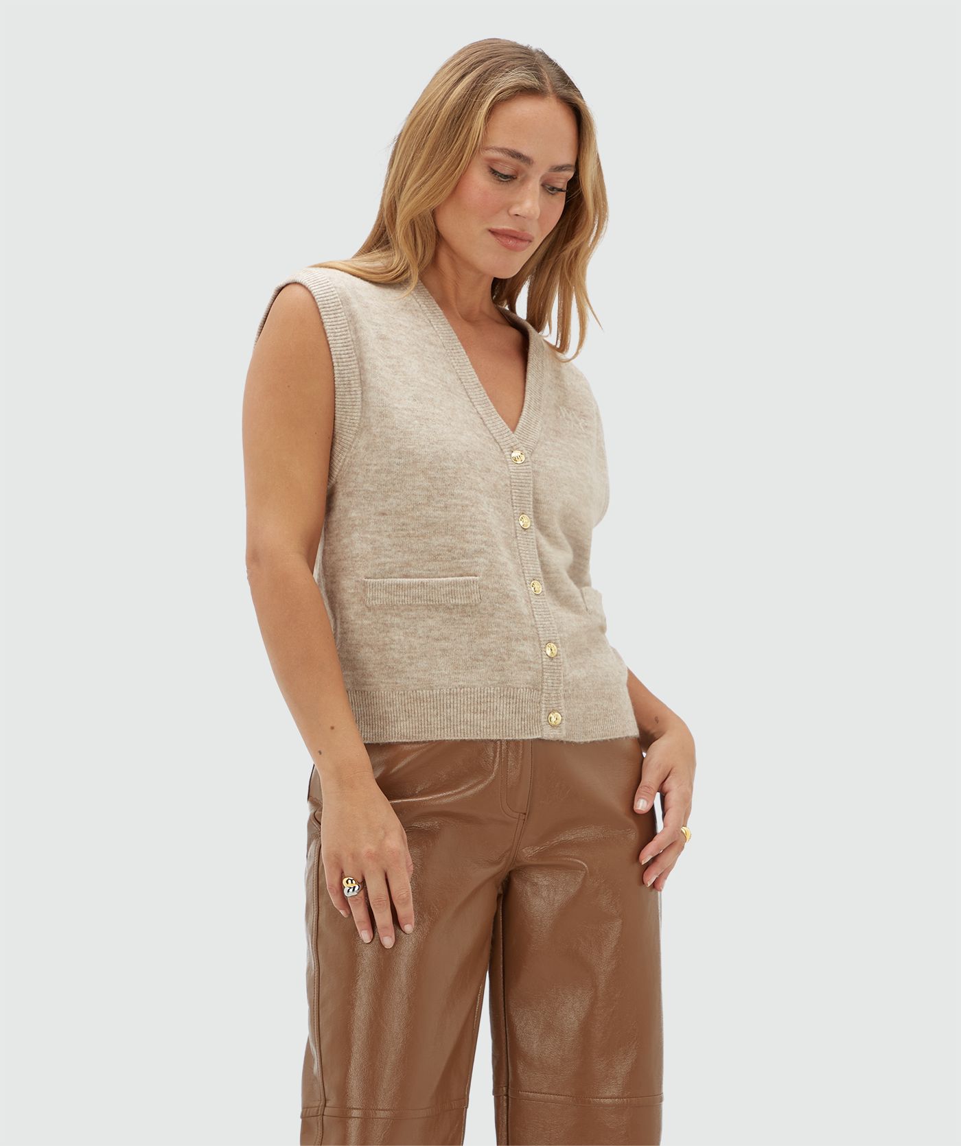 Model draagt een beige gilet met gouden knoopjes en een JOSH V logo op de borst. Het gilet is gecombineerd een bruine lak look broek.