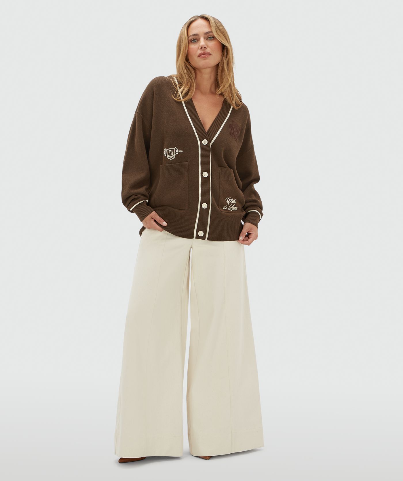 Model draagt een oversized donkerbruin vest met gebroken witte details. Het vest heeft ook twee steekzakken aan de voorkant. 