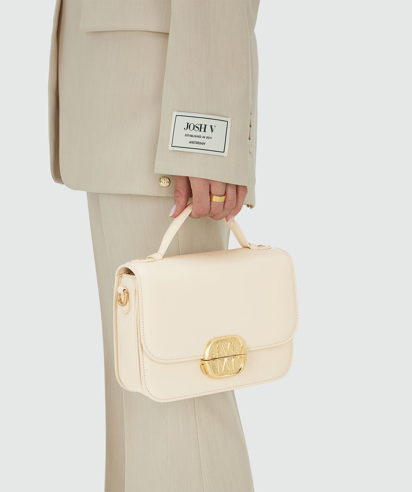 Het model houdt een tas in Crème White met een goudkleurige bucklesluiting vast aan het handhengsel