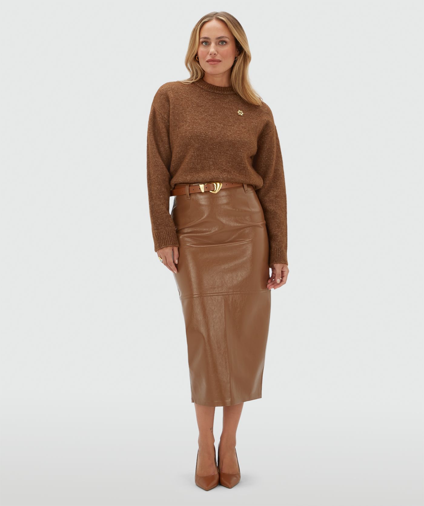 Model draagt een bruine trui met een klein gouden JOSH V logo op de borst. De trui is gecombineerd met een bruine lange lak look rok.