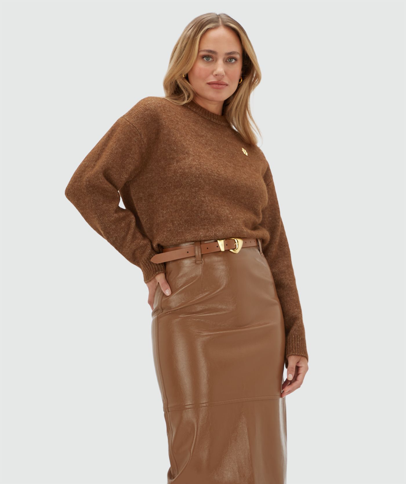 Model draagt een bruine trui met een klein gouden JOSH V logo op de borst. De trui is gecombineerd met een bruine lange lak look rok.