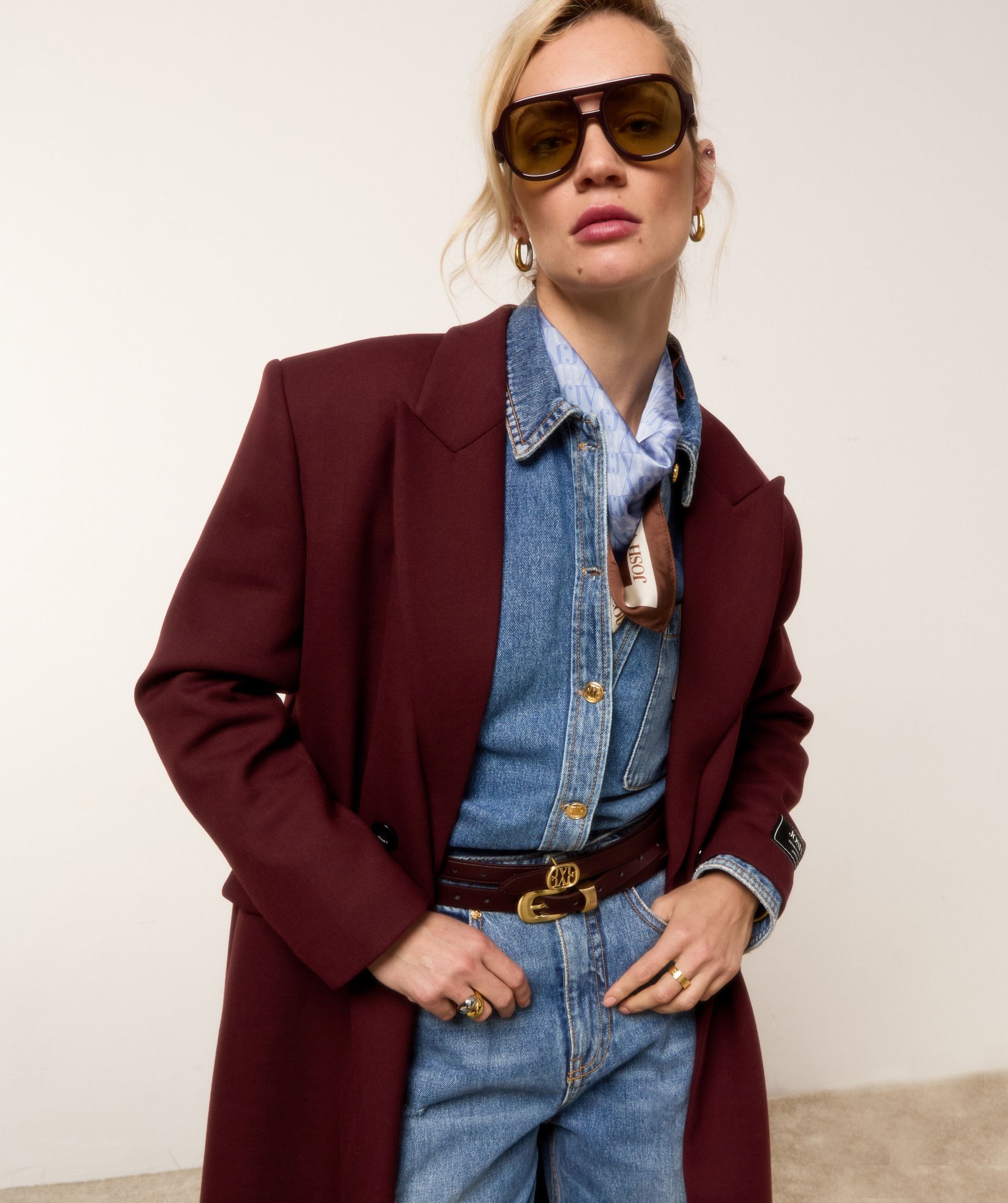 Model draagt een spijkerblouse en een spijkerbroek met een donkerrode coat en de donkerrode zonnebril.