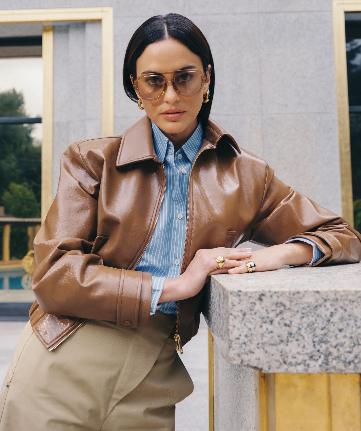 Model leunt tegen een muur en draagt een beige zonnebril met een beige pantalon en een blauw met wit gestreepte blouse met een lak look jasje.