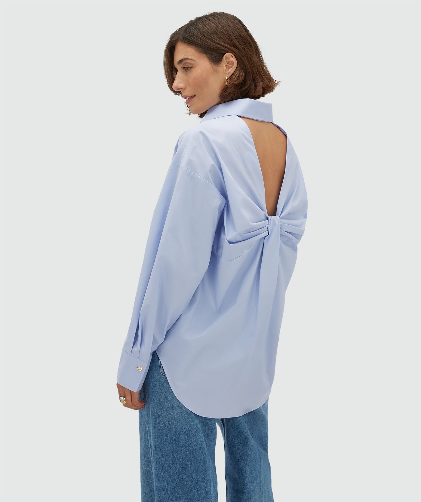 Zijaanzicht van een model in een oversized licht blauwe blouse met strikdetail aan de achterzijde. 