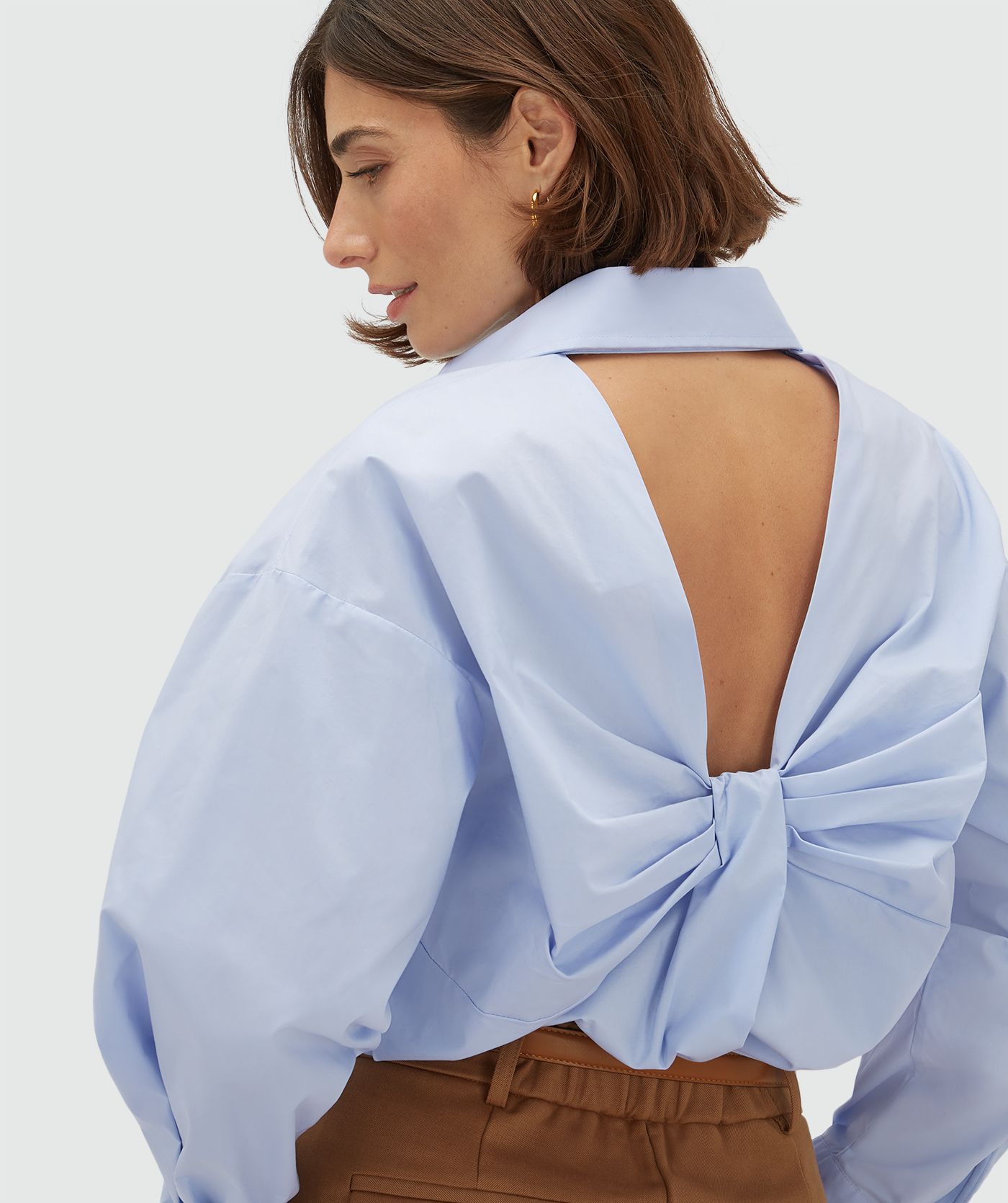 Zijaanzicht van een model in een oversized licht blauwe blouse met strikdetail aan de achterzijde. 