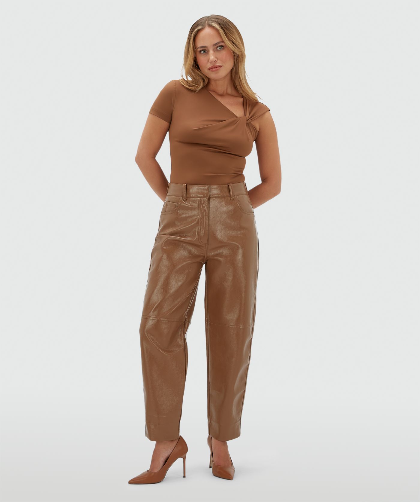 Model draagt een lichtbruine body met een knoop detail op de borst en is gecombineerd met een lichtbruine lak look broek.
