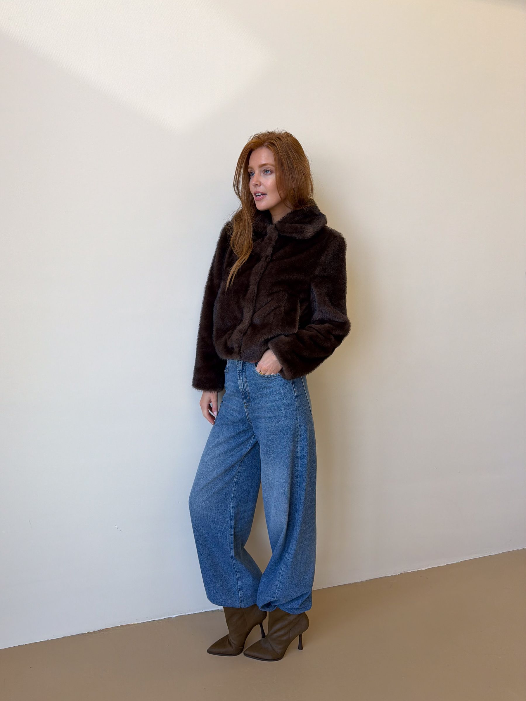 Model draagt een donkerbruine bond jas gecombineerd met een jeans en donkergroene laarsjes.