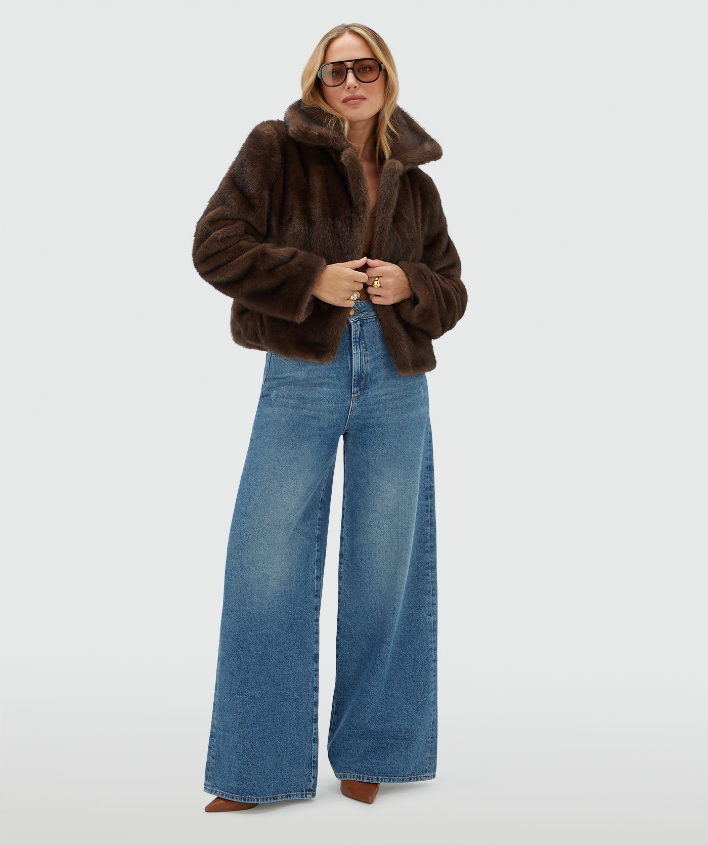 Model draagt een donkerbruine bond jas gecombineerd met een jeans en een donkerbruine zonnebril.