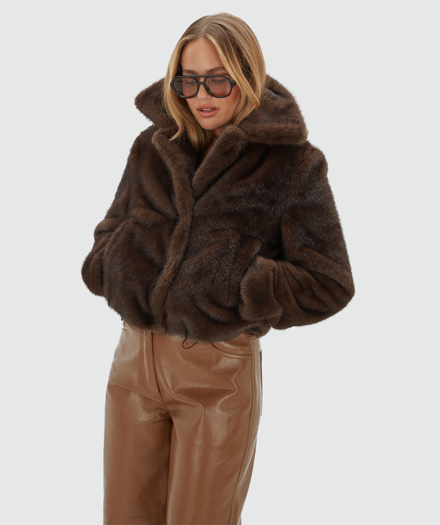 Model draagt een donkerbruine bond jas met een lichtbruine lak look broek.