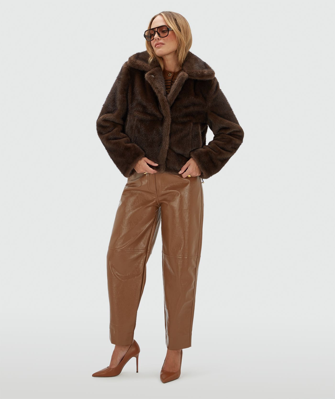 Model draagt een donkerbruine bond jas met een lichtbruine lak look broek.