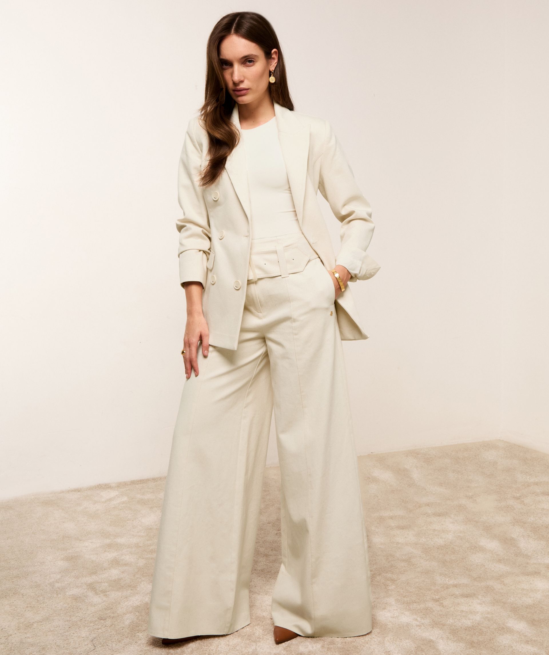 Model staat bij een witte muur en draagt de gebroken witte wide leg jeans met de bijpassende blazer.