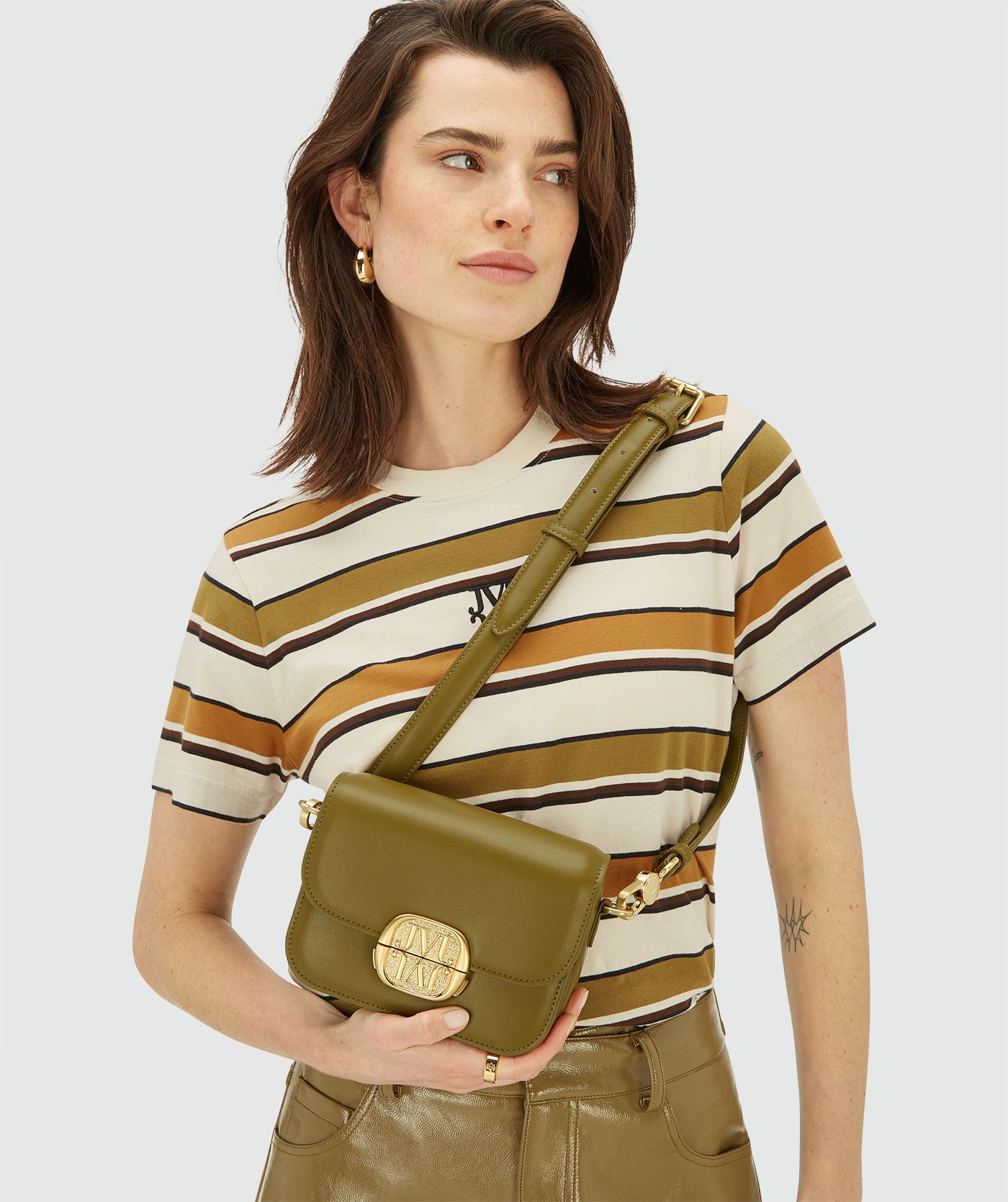 Model draagt een Olive Green schoudertas met klepsluiting en goudkleurig logo-embleem aan de voorzijde. De tas heeft een verstelbare schouderband met goudkleurige details