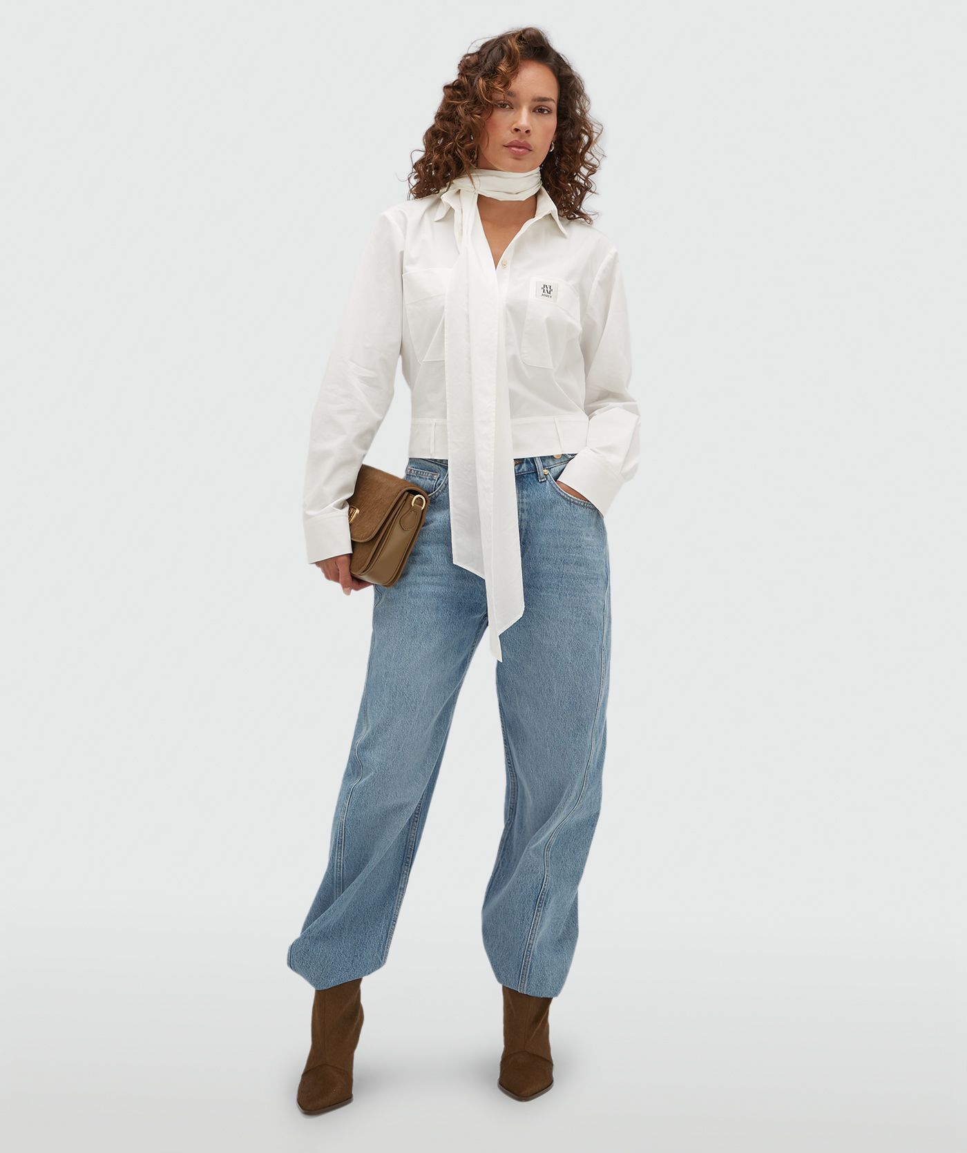 Model draagt een witte blouse met een strap die je op verschillende manieren kan dragen en is voorzien van een borstzak met een JOSH-V-label.