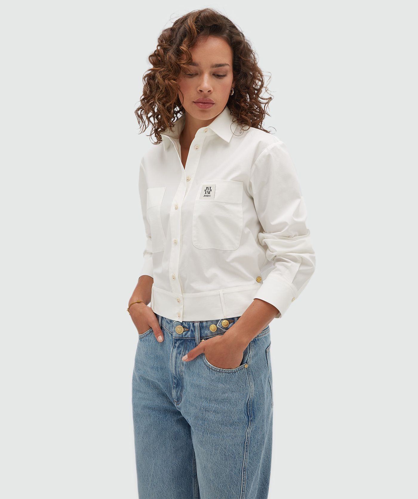 Model draagt een witte blouse met een strap die je op verschillende manieren kan dragen en is voorzien van een borstzak met een JOSH-V-label.