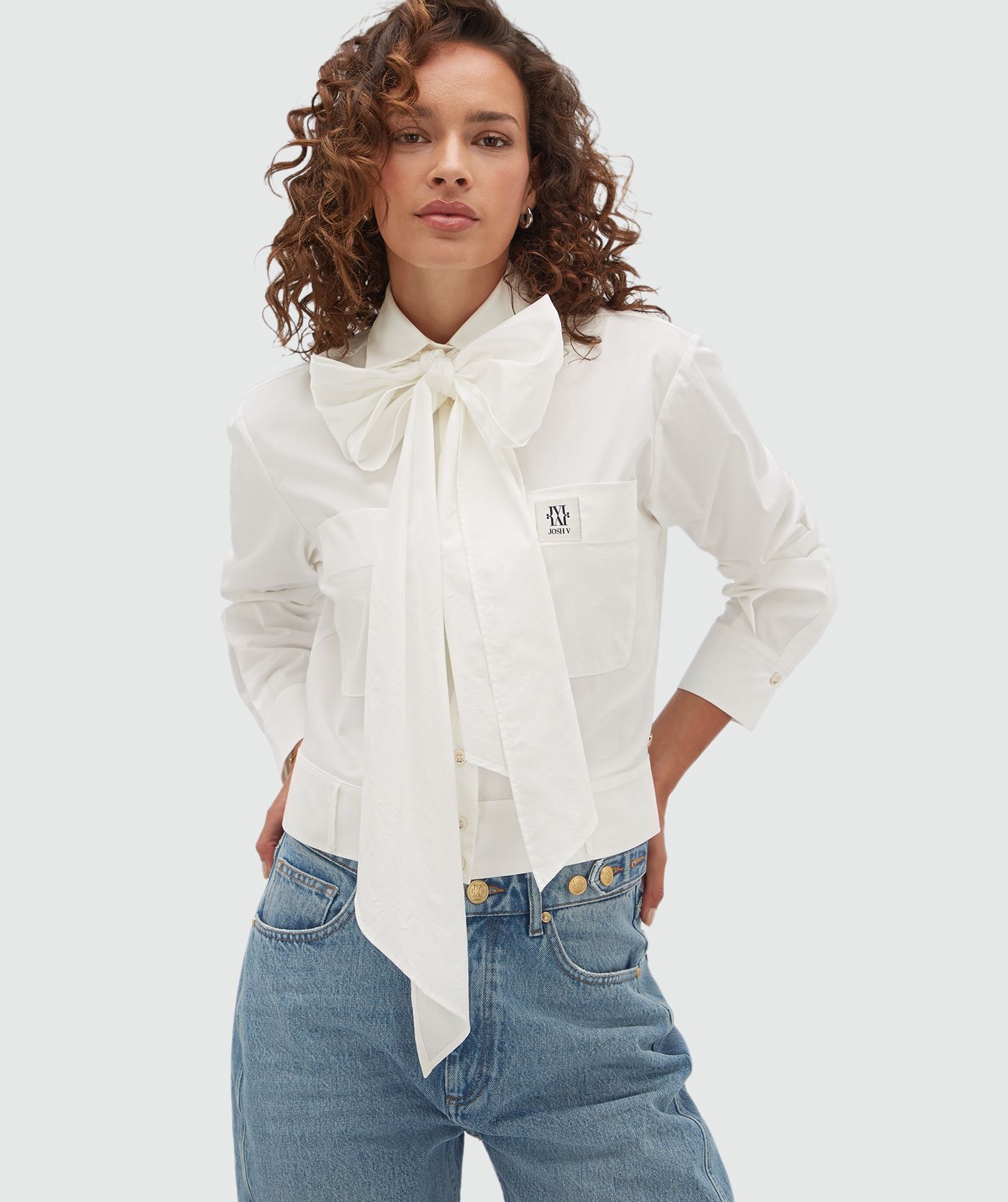 Model draagt een witte blouse met een strap die je op verschillende manieren kan dragen en is voorzien van een borstzak met een JOSH-V-label.