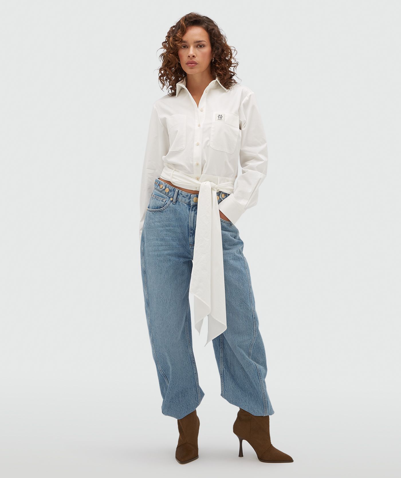 Model draagt een witte blouse met een strap die je op verschillende manieren kan dragen en is voorzien van een borstzak met een JOSH-V-label.