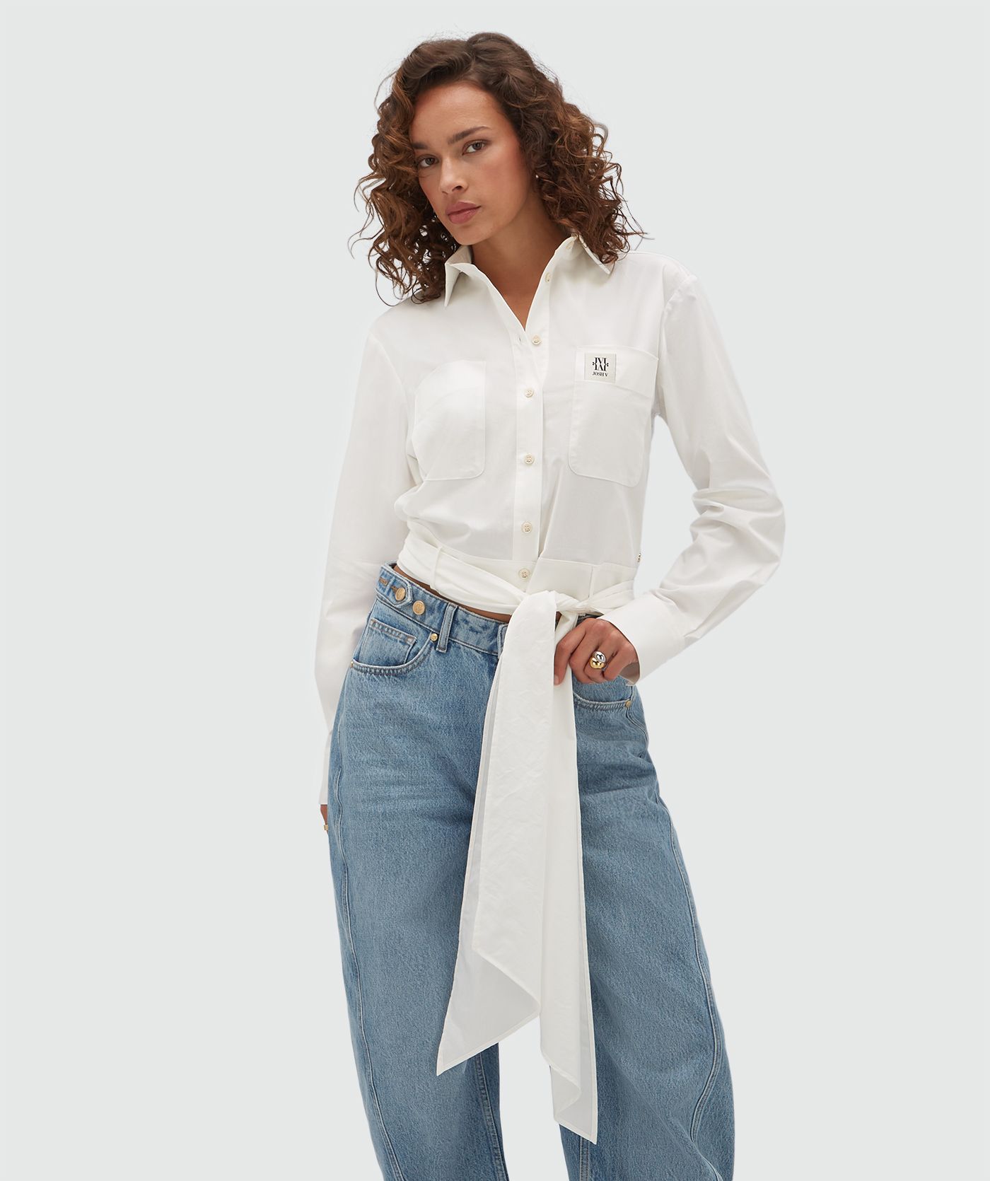 Model draagt een witte blouse met een strap die je op verschillende manieren kan dragen en is voorzien van een borstzak met een JOSH-V-label.