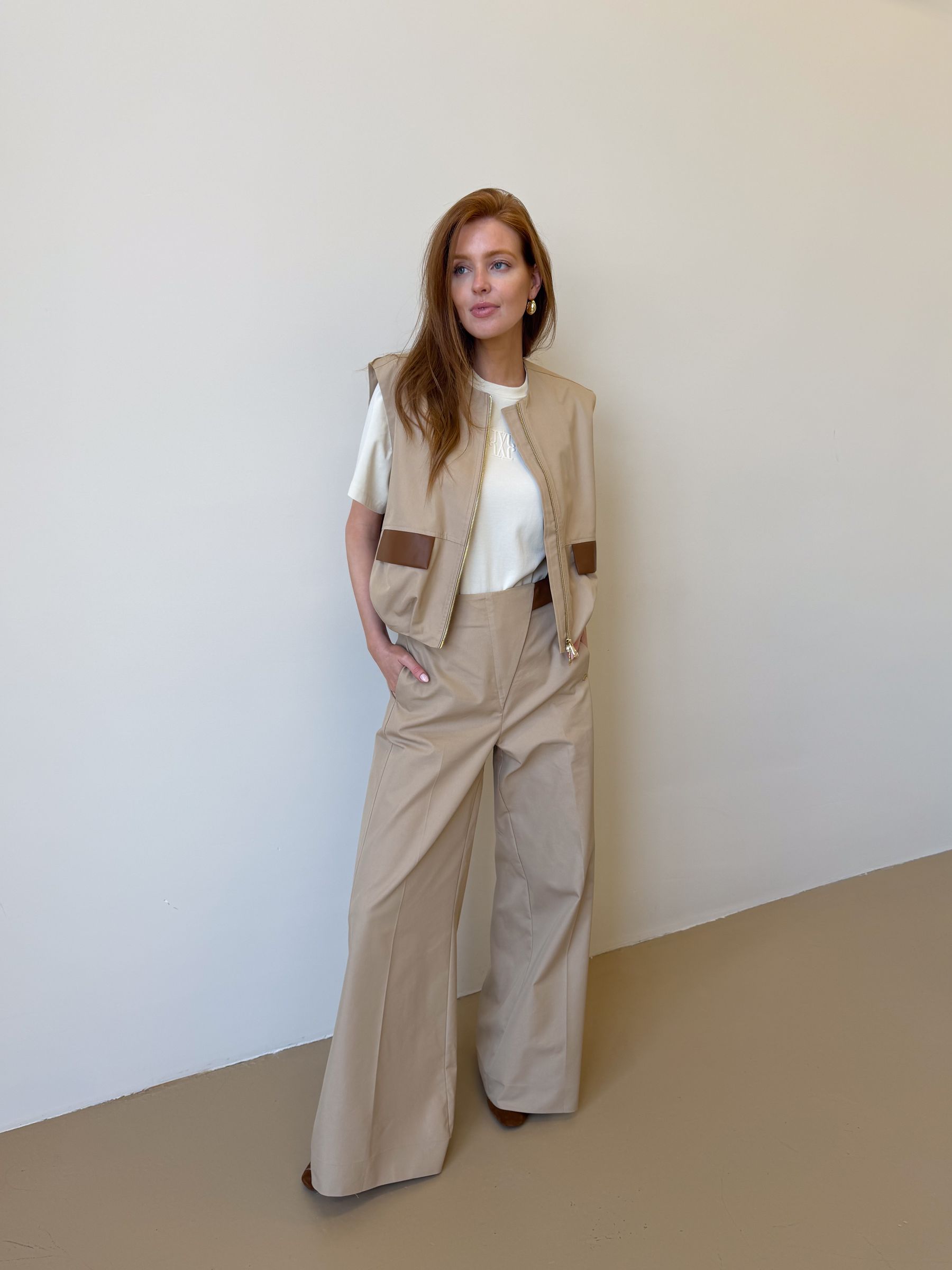 Model draagt een beige gilet met een rits en leren details bij de zakken op de voorkant. Het gilet is gecombineerd met wit T-shirt en de bijpassende broek.