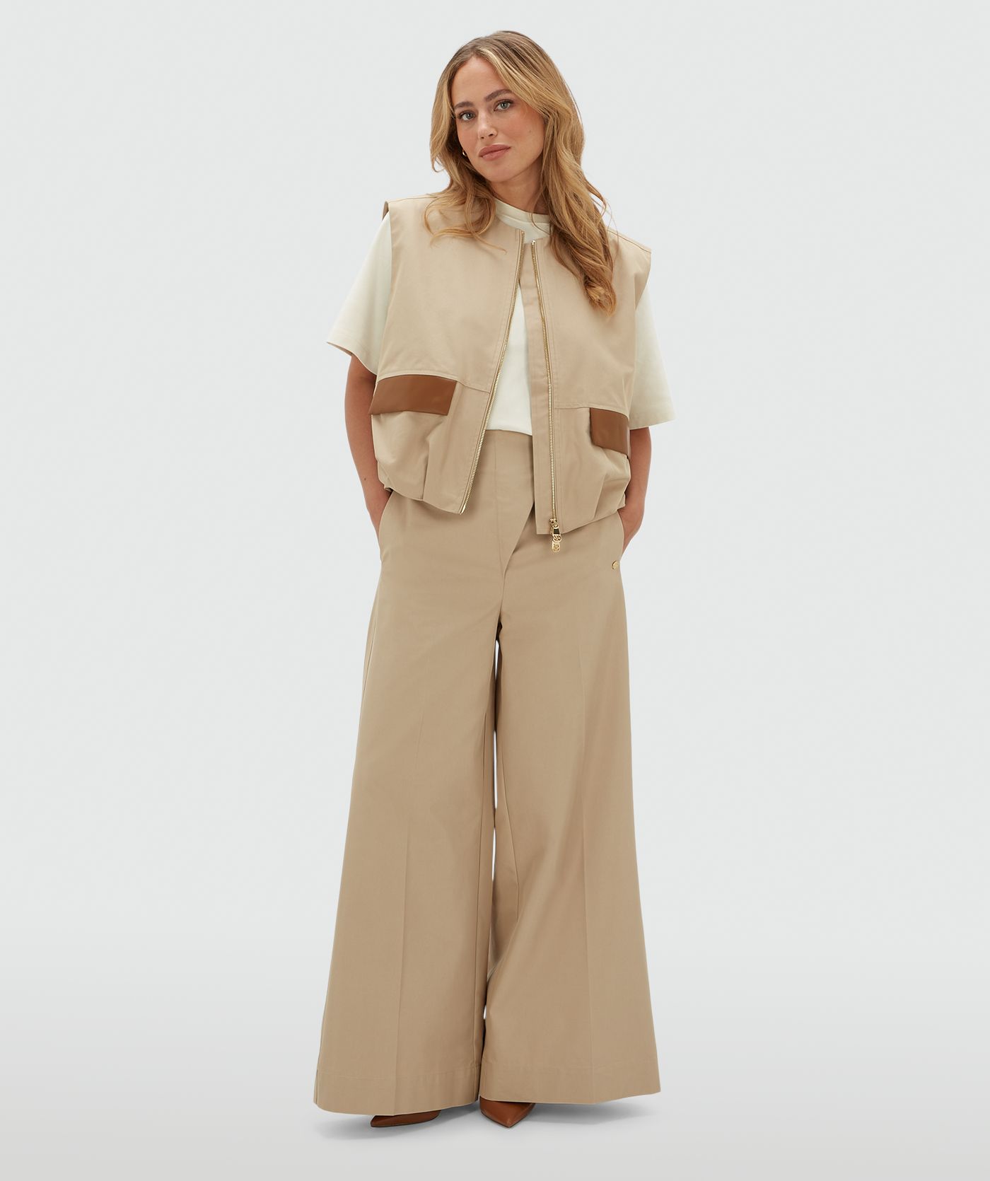 Model draagt een beige gilet met een rits en leren details bij de zakken op de voorkant. Het gilet is gecombineerd met wit T-shirt en de bijpassende broek.