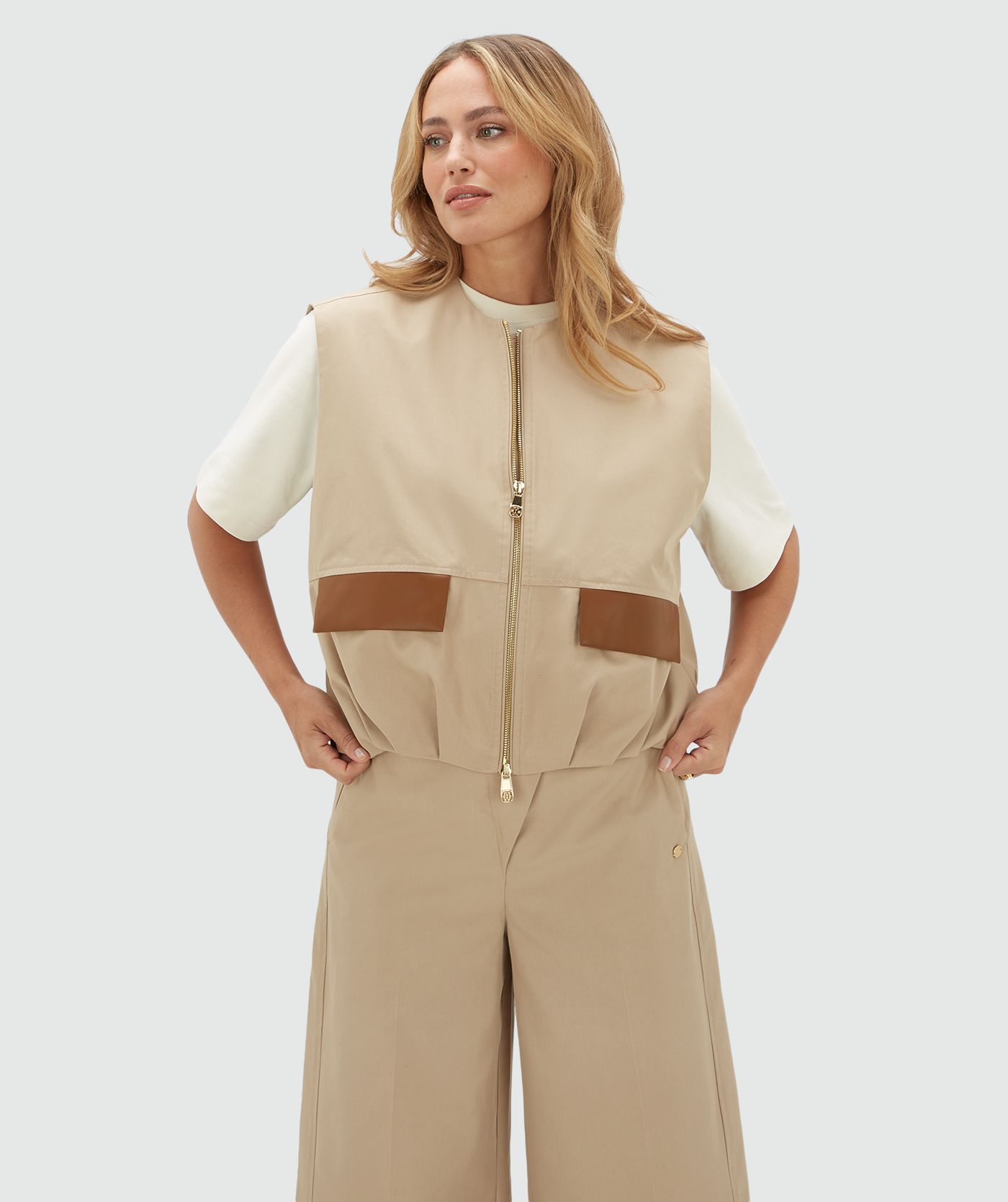 Model draagt een beige gilet met een rits en leren details bij de zakken op de voorkant. Het gilet is gecombineerd met wit T-shirt en de bijpassende broek.