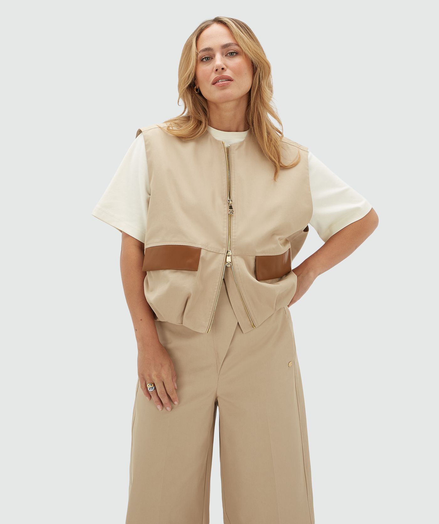Model draagt een beige gilet met een rits en leren details bij de zakken op de voorkant. Het gilet is gecombineerd met wit T-shirt en de bijpassende broek.