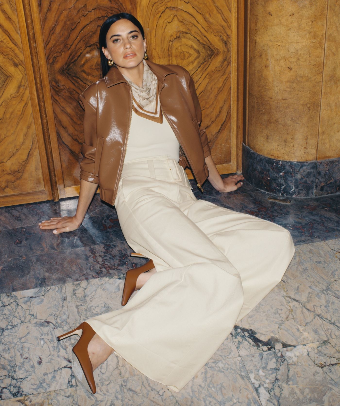 Model zit op de grond en heeft een beige outfit aan met daaroverheen het bruine lak look jasje.