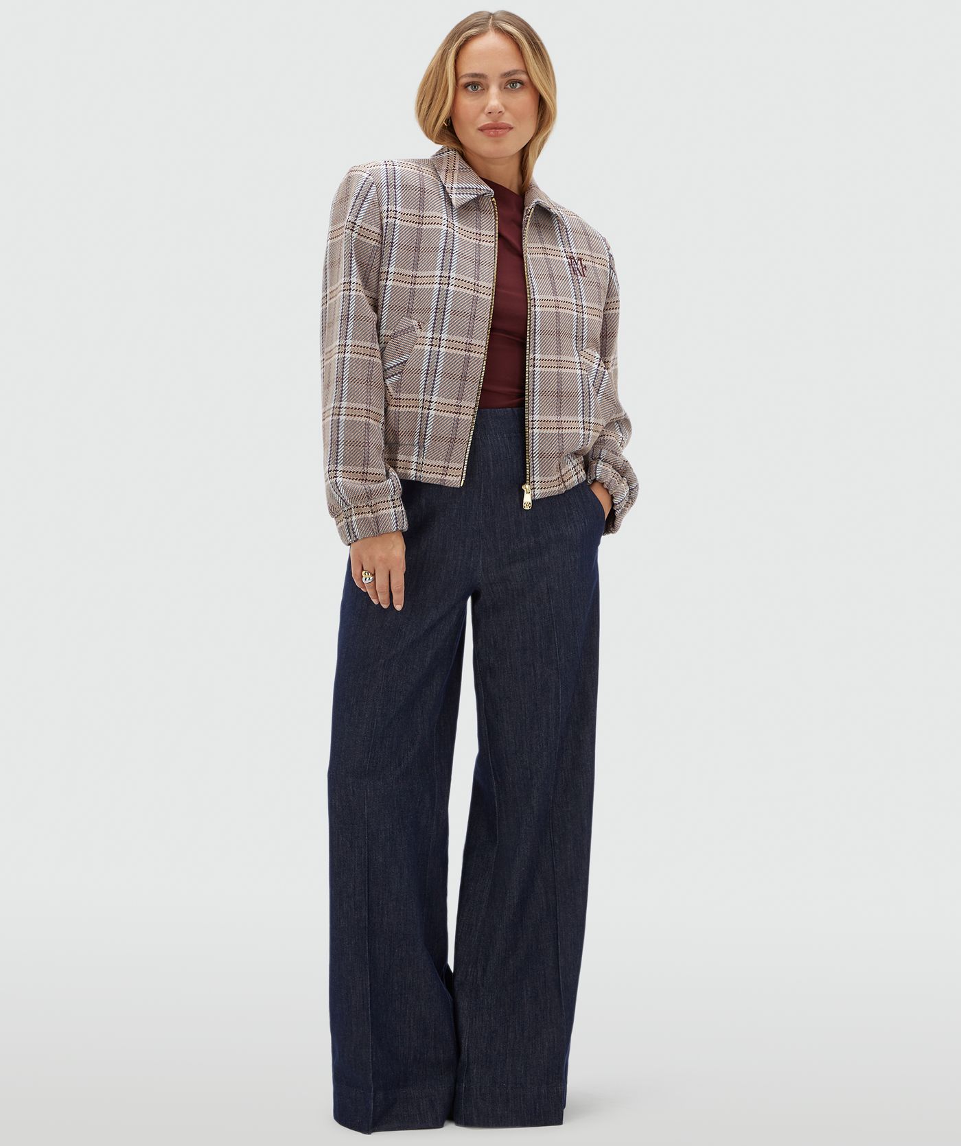 Model draagt een beige, bruin en donkerrood geruit jasje met rits gecombineerd met een donkerblauwe pantalon en een rood topje.